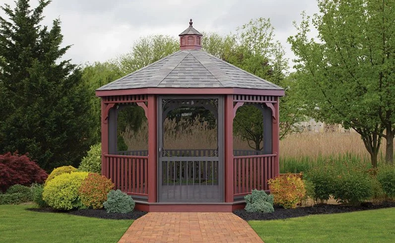 A round, open-sided gazebo with a shingled roof and decorative trim, situated on a brick pathway surrounded by landscaping with bushes and trees.
