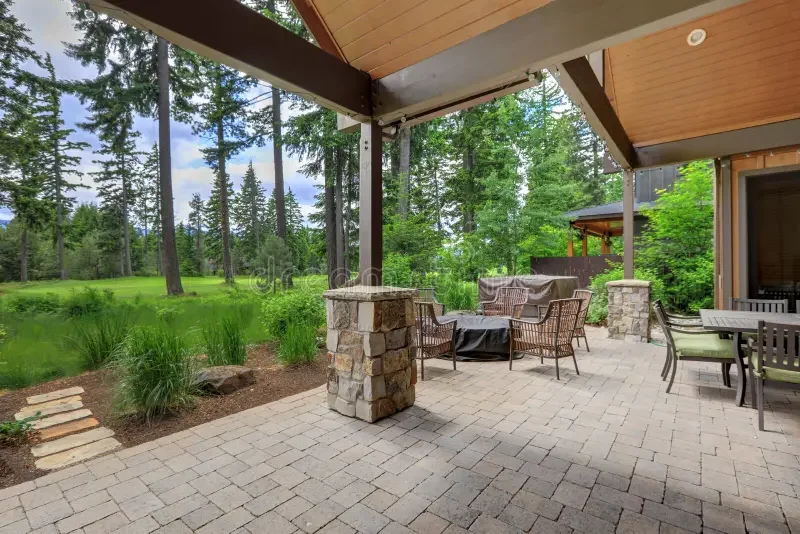 Covered outdoor patio with stone pillars, wicker chairs, and a dining table, overlooking a lush green forest and grass yard.