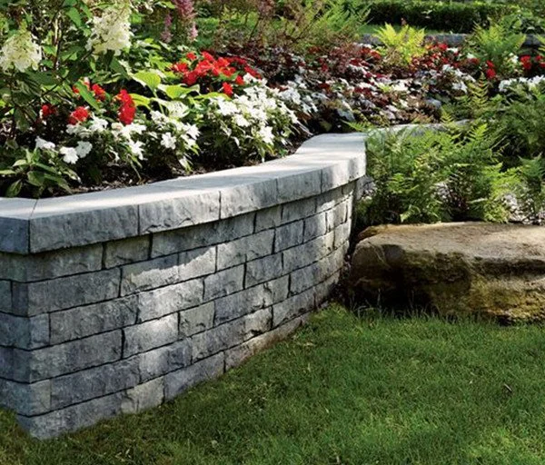 A stone garden wall with colorful flowers and green plants behind it, and a large rock to the right, on a grassy lawn.