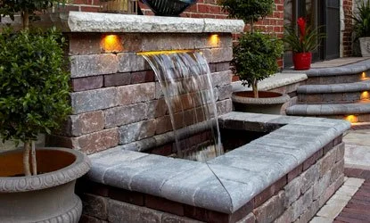 A brick water fountain with water flowing from a spout into a rectangular basin, surrounded by potted plants and steps with embedded lighting.
