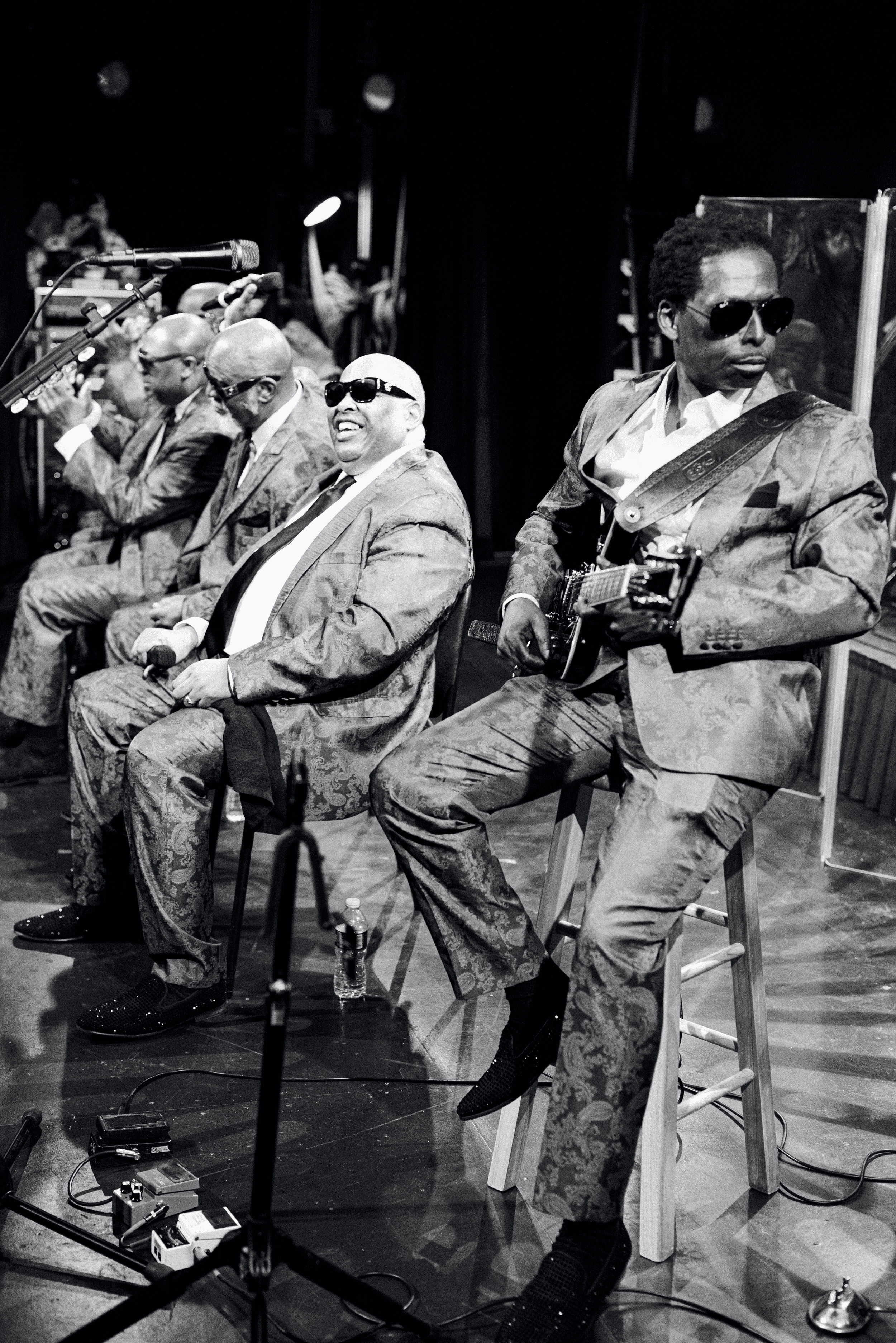 A black and white photo of a music band performing on stage, with musicians wearing patterned suits and sunglasses, one playing guitar and others seated with microphones and music equipment.