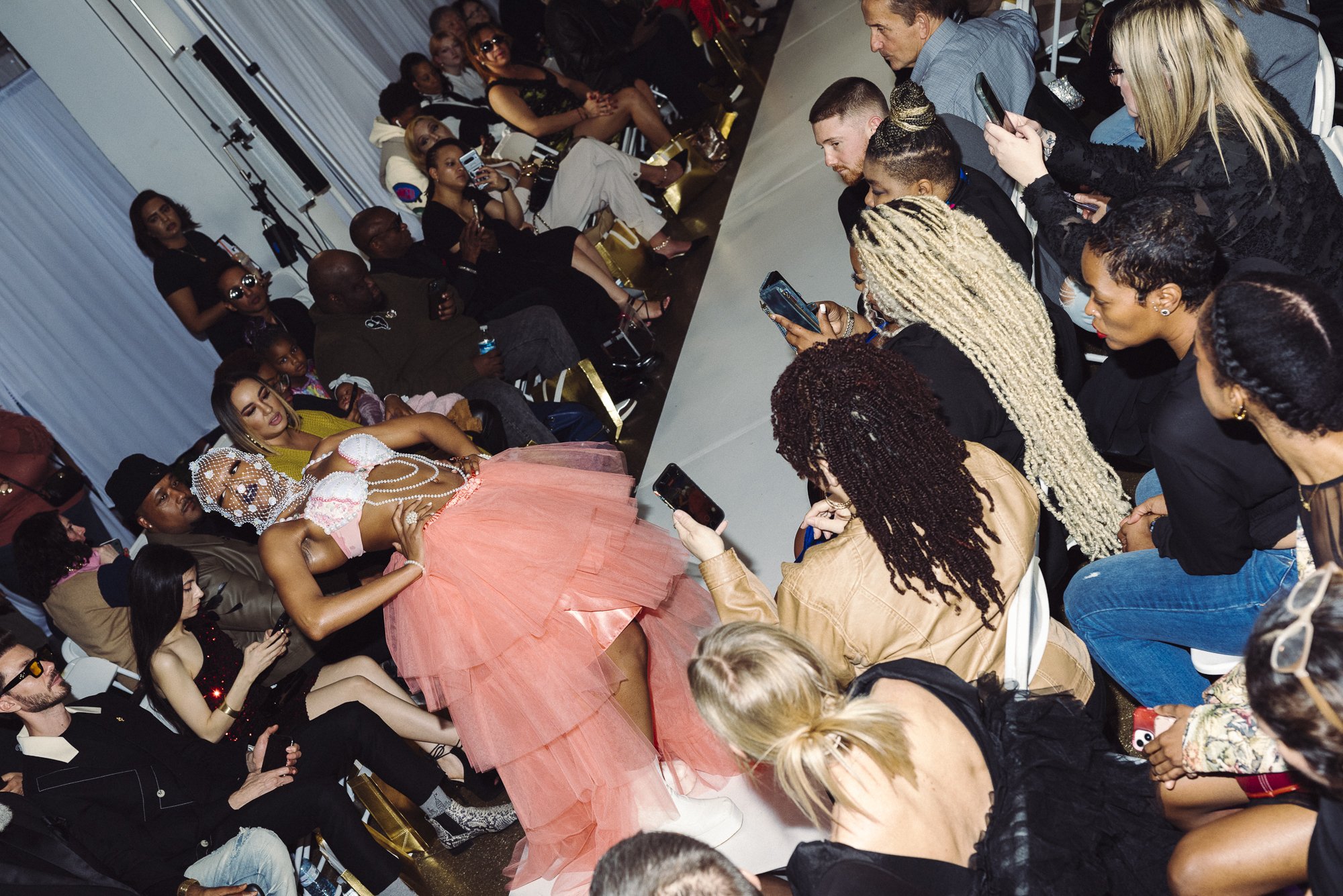 Fashion runway with model wearing a pink tulle skirt and a top with pearl and fabric embellishments, surrounded by an attentive audience taking photos and videos.