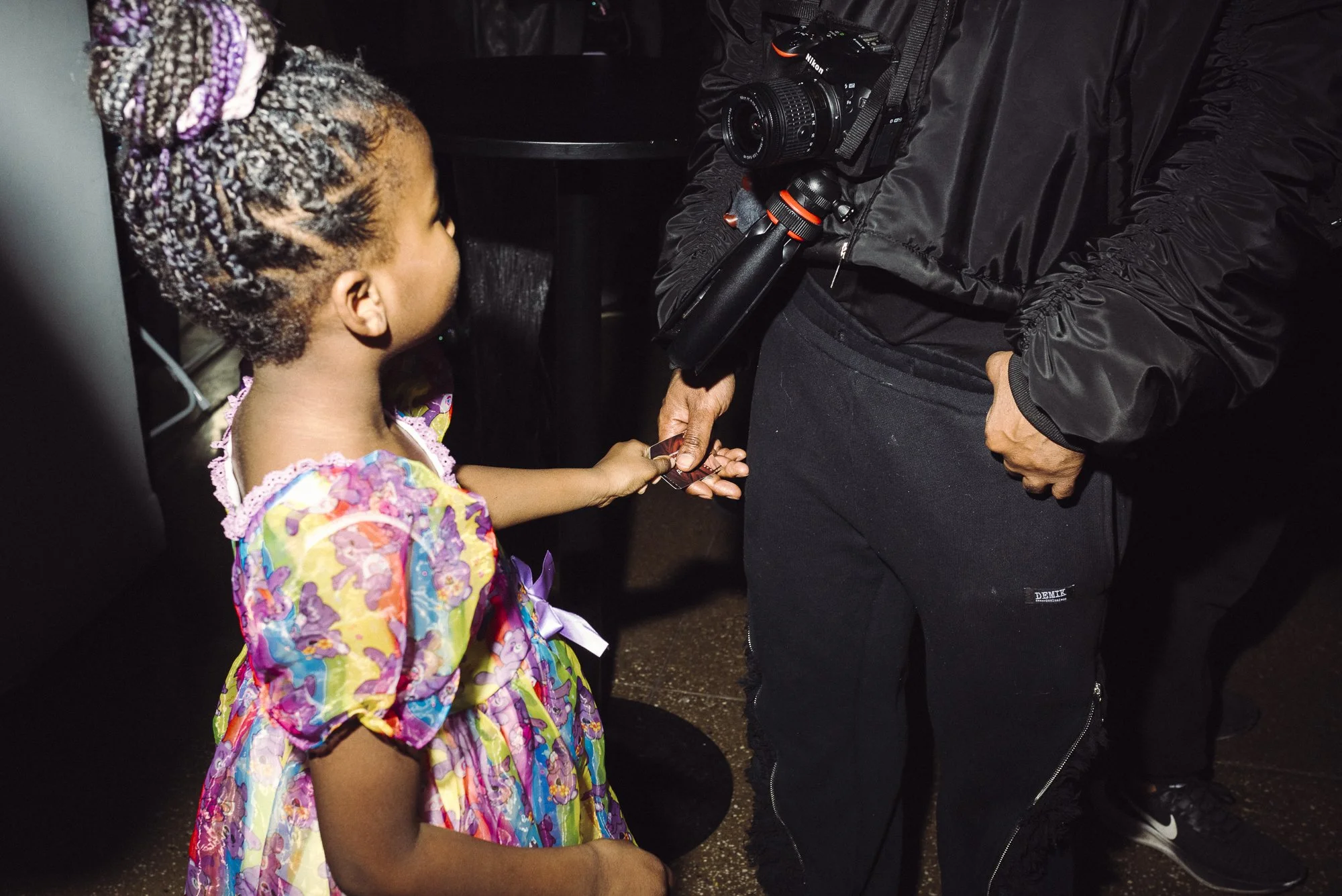 A young girl with braided hair and wearing a colorful dress is handing over a key to an adult dressed in black, who is holding a camera.