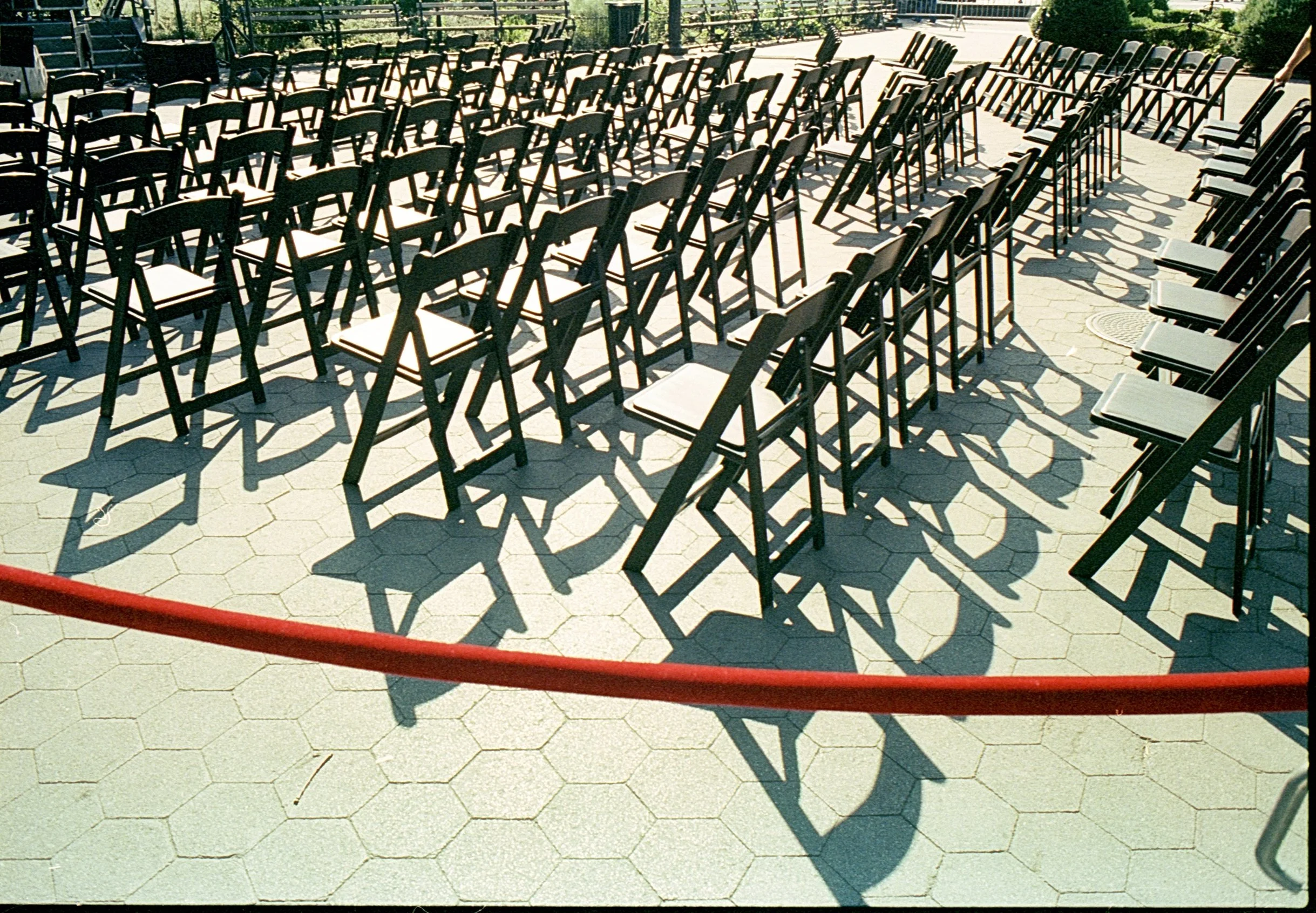 Outdoor setting with rows of black folding chairs on hexagonal pavers, separated by a red barrier.