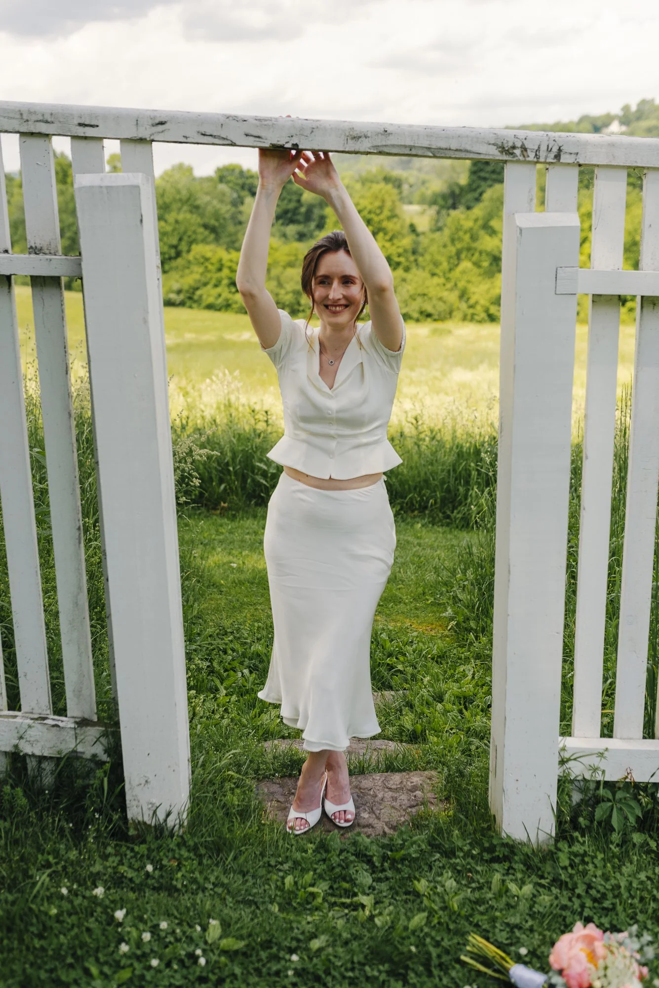 Woman in white dress standing outdoors at a gate, smiling, with greenery and trees in the background.