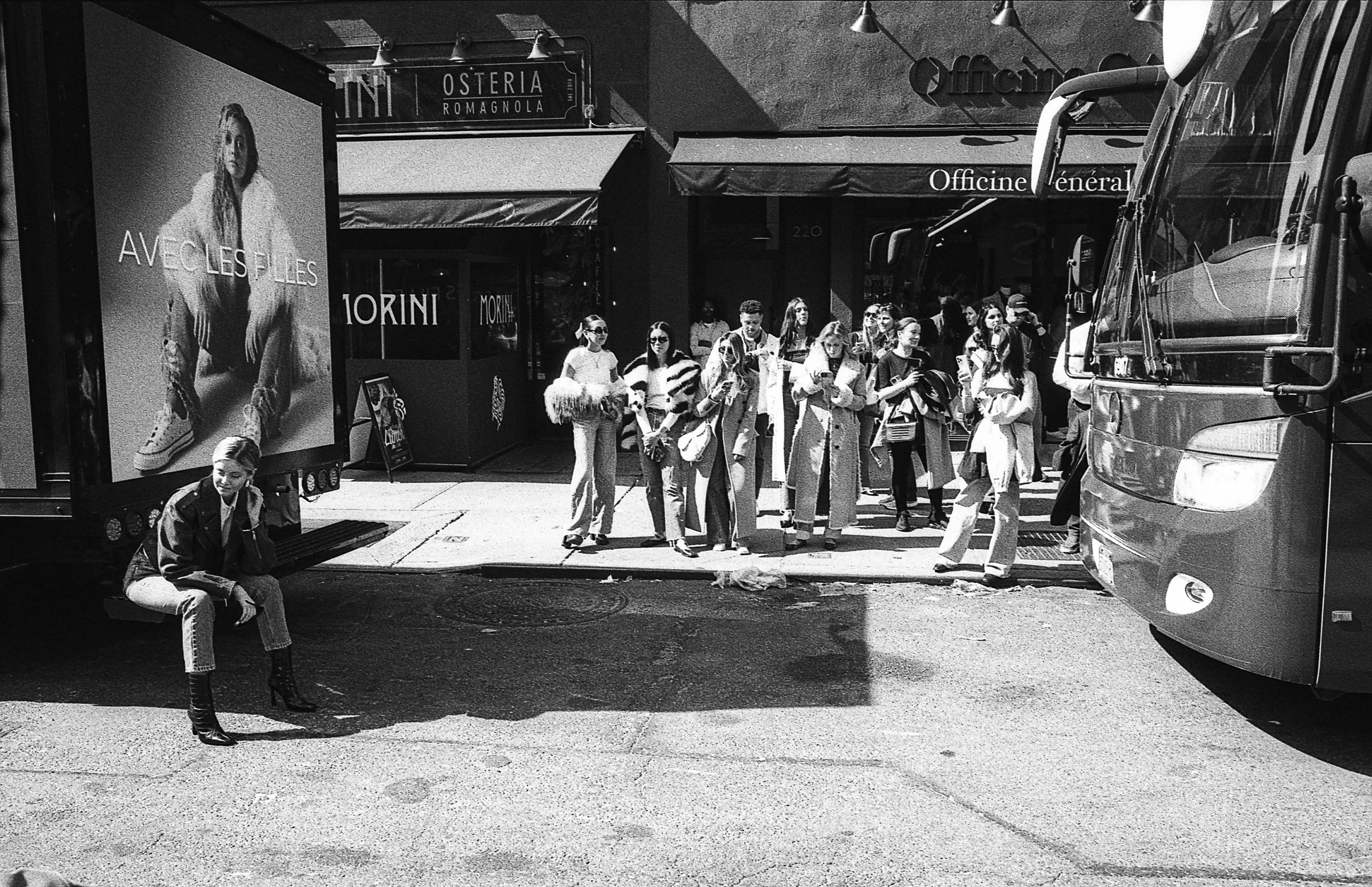 Black and white street scene with a woman sitting near a fashion advertisement on a truck, and a group of people standing on a sidewalk. A bus is parked nearby, and storefronts are visible in the background.
