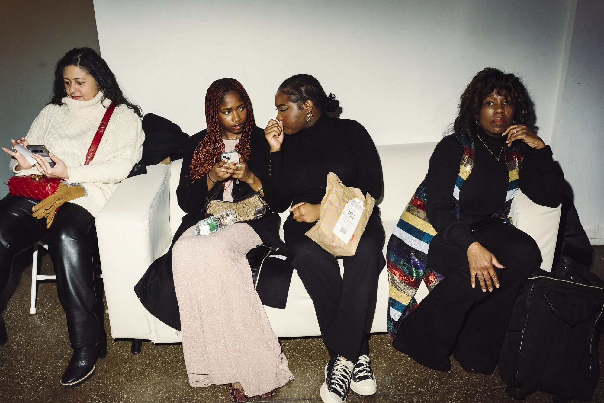 Four women sitting on a white sofa, two using their phones and one looking at her phone, with a water bottle and shopping bag nearby. The woman on the far right is sitting with a large black bag beside her.