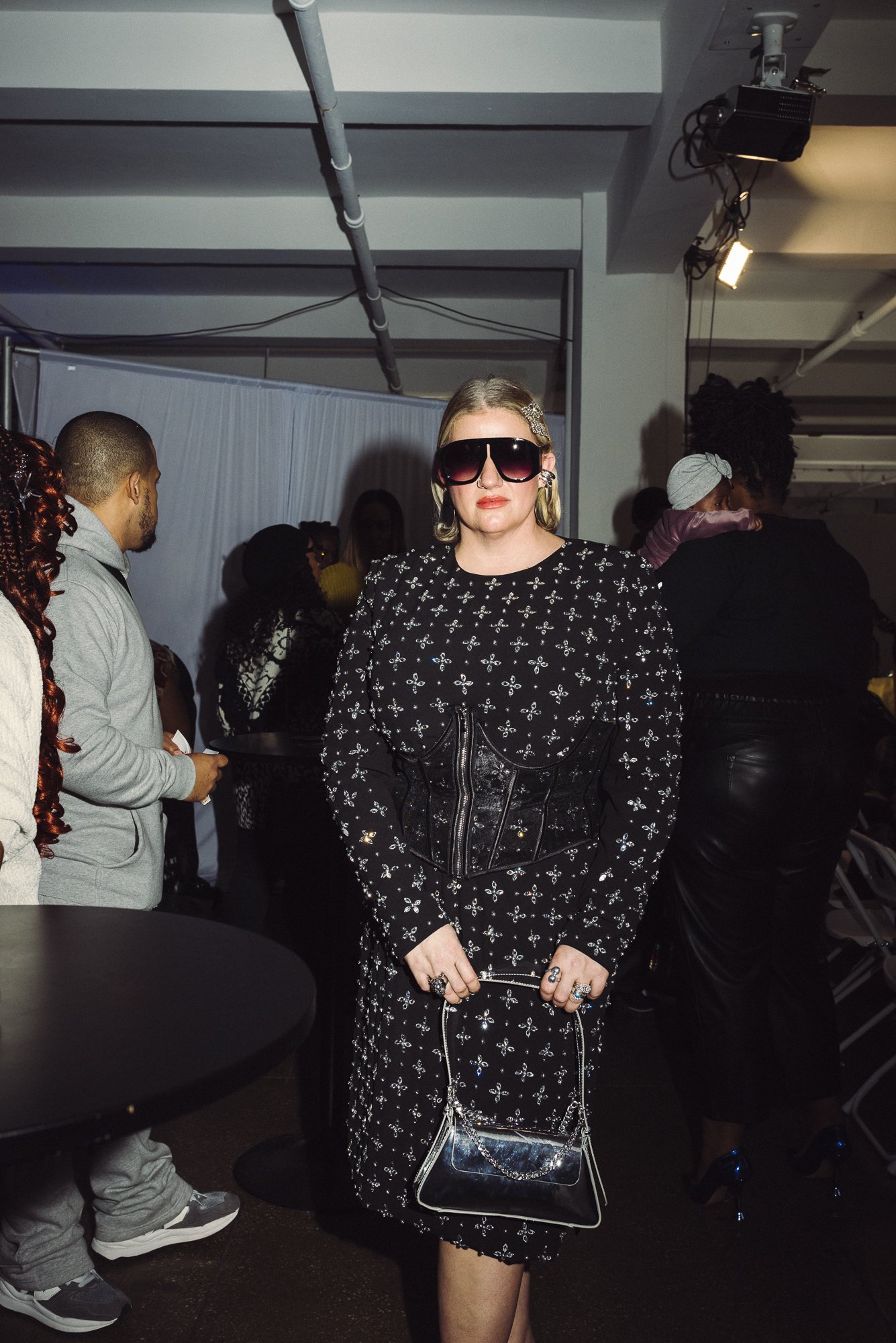 Fashionably dressed woman wearing large sunglasses and black, patterned dress with matching handbag at social gathering.