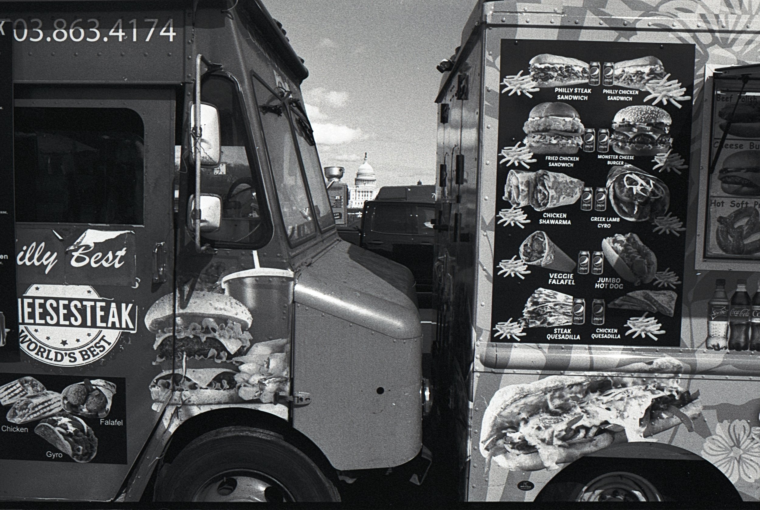 Two food trucks parked side by side with images of various sandwiches and wraps displayed on their exteriors. The U.S. Capitol building is visible in the background.