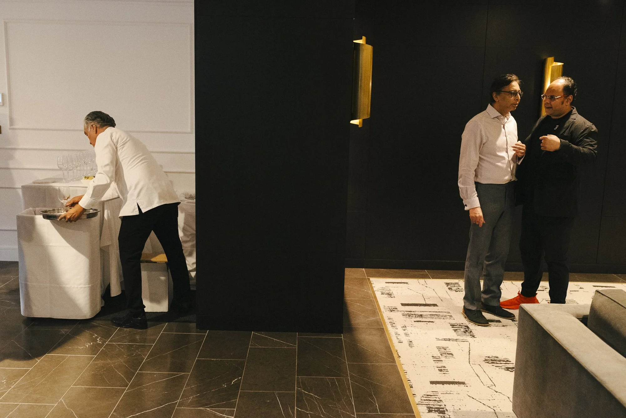 Three men at an indoor gathering; one man in white serving food at a table on the left, and two men on the right engaged in conversation against a dark wall, one wearing a checkered shirt and the other in black with red shoes.