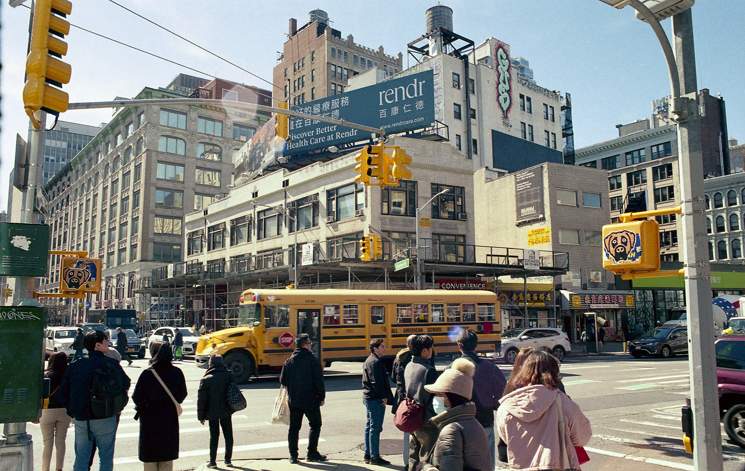 Urban street scene with pedestrians crossing, yellow school bus, and various storefronts. Prominent billboards and traffic lights are visible against a backdrop of multi-story buildings.