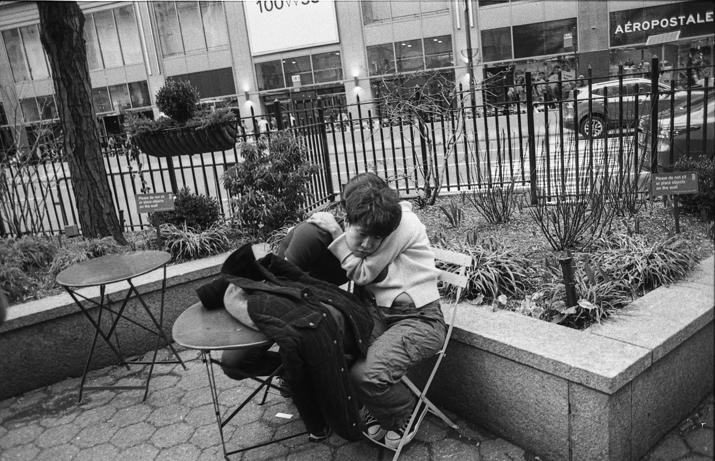 Black and white photo of a person sitting alone at a small outdoor table. They are leaning forward with their head on the table, appearing contemplative. A jacket is draped over the table. The setting is an urban park area, with a fence and a retail 