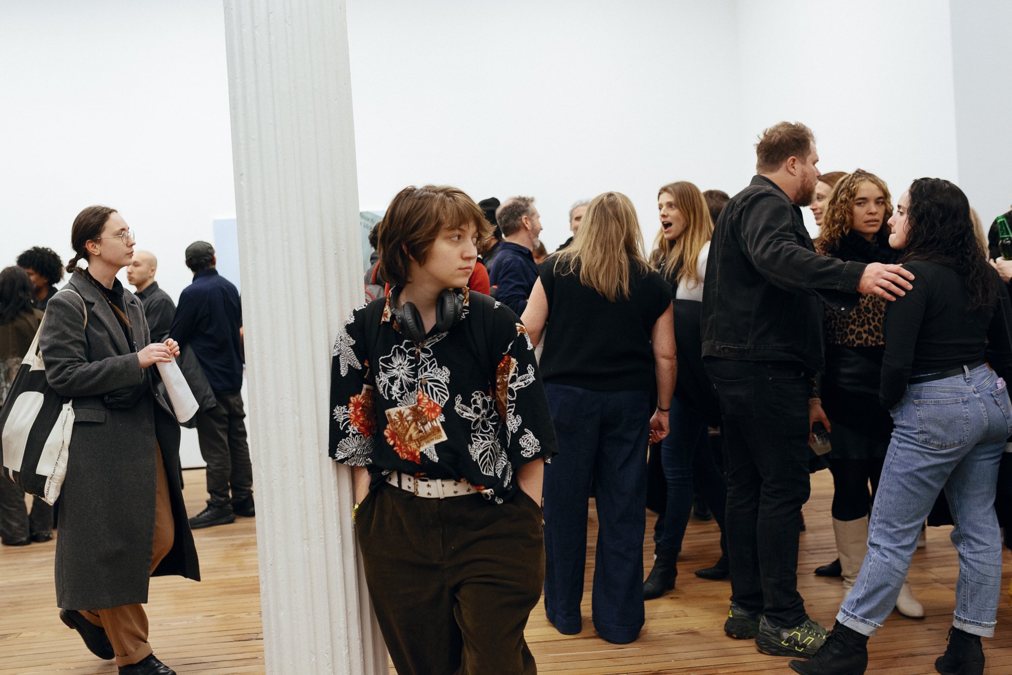 A crowded art gallery with people socializing. A person in a floral shirt leans against a column, wearing headphones. Others are engaged in conversation around the room. The gallery has wooden floors and white walls.