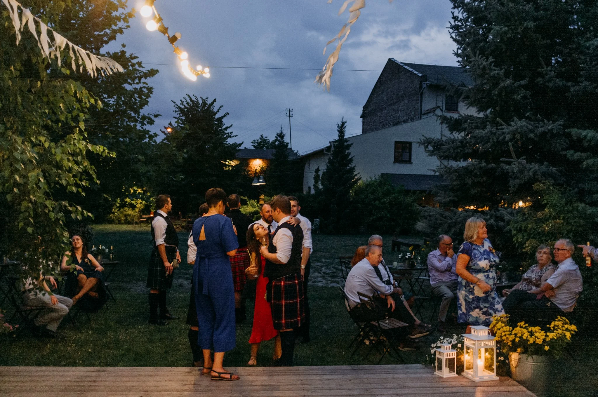 People socializing and dancing outdoors at dusk, with string lights and lanterns, surrounded by trees and a rustic building in the background.