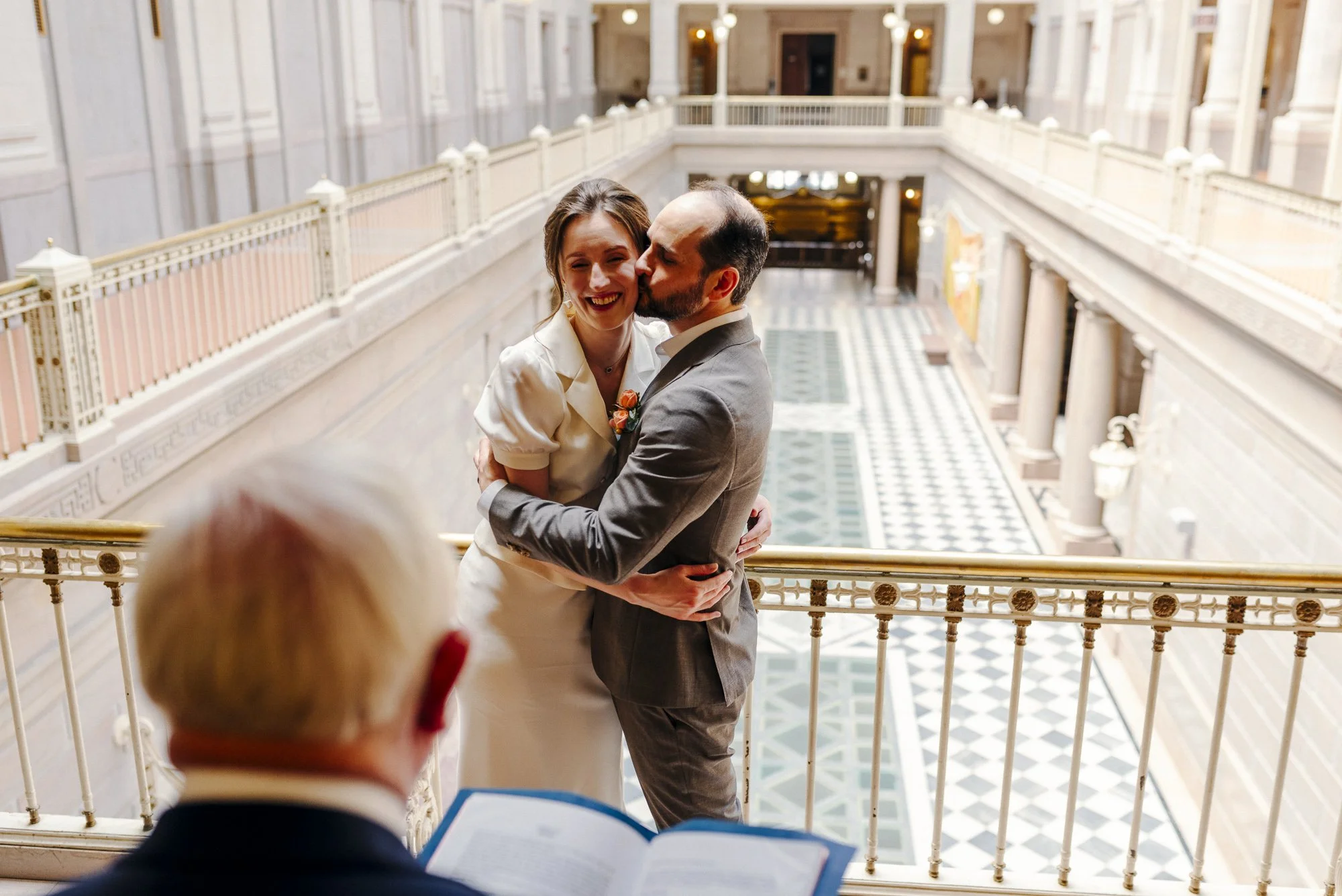 A bride and groom hugging and smiling at a wedding ceremony or reception inside a grand, historic building with ornate railings and high ceilings, while an officiant or celebrating guest reads from a book.