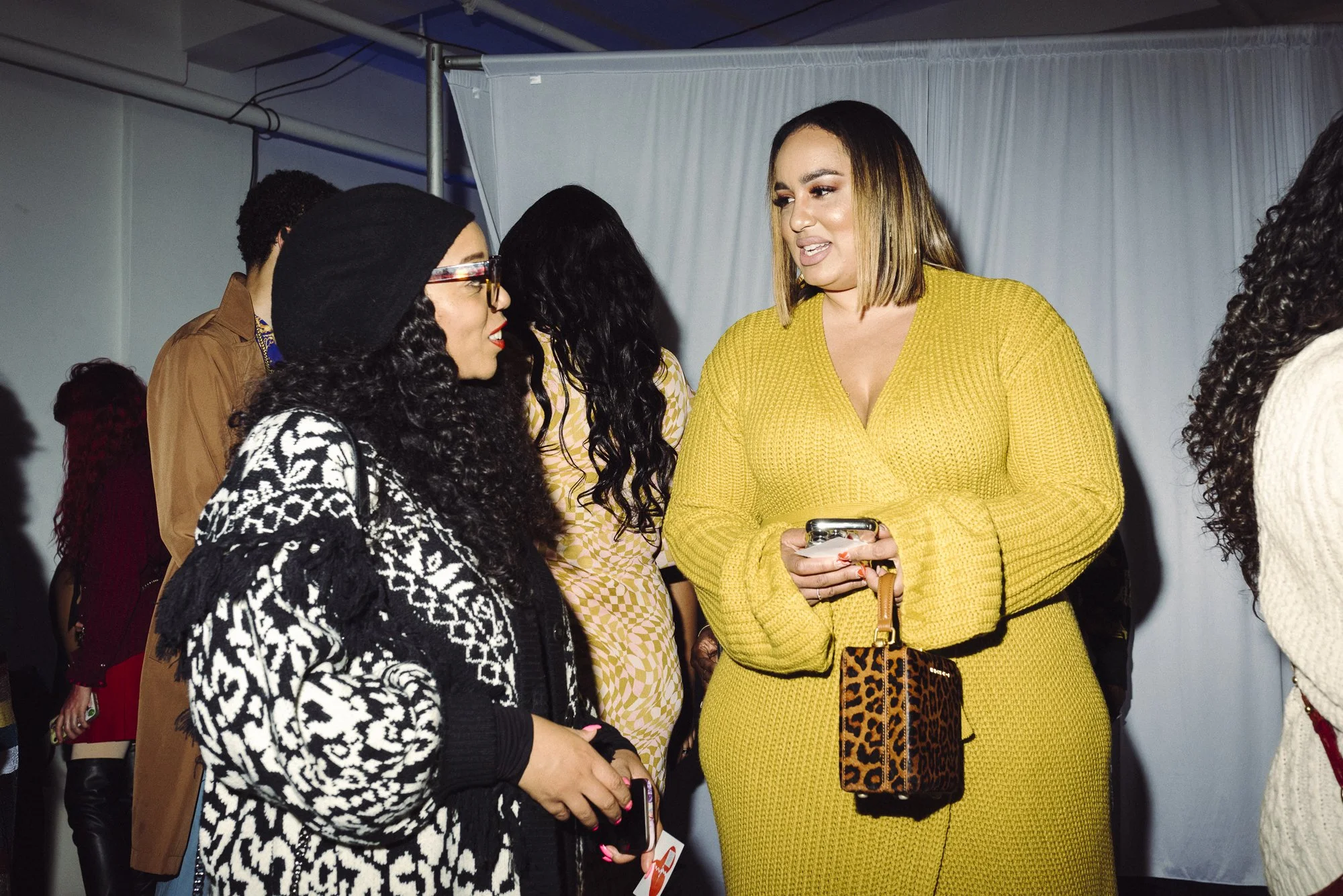 Two women in conversation at an indoor event. The woman on the right wears a yellow dress and holds a phone and a leopard-print purse. The woman on the left wears glasses, a black hat, and a black and white patterned sweater.