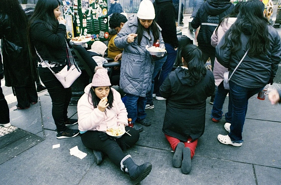 Group of people eating near a street vendor in a crowded outdoor area. One person is sitting on the ground eating from a plate, while others are standing and snacking. Winter clothing and street food visible.