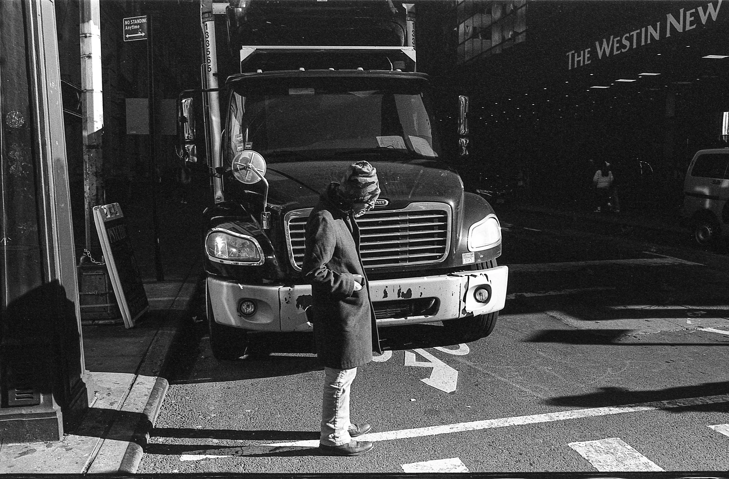 Black and white photo of a person standing in front of a large truck on a city street, wearing a coat and hat. "The Westin New York" is visible in the background.