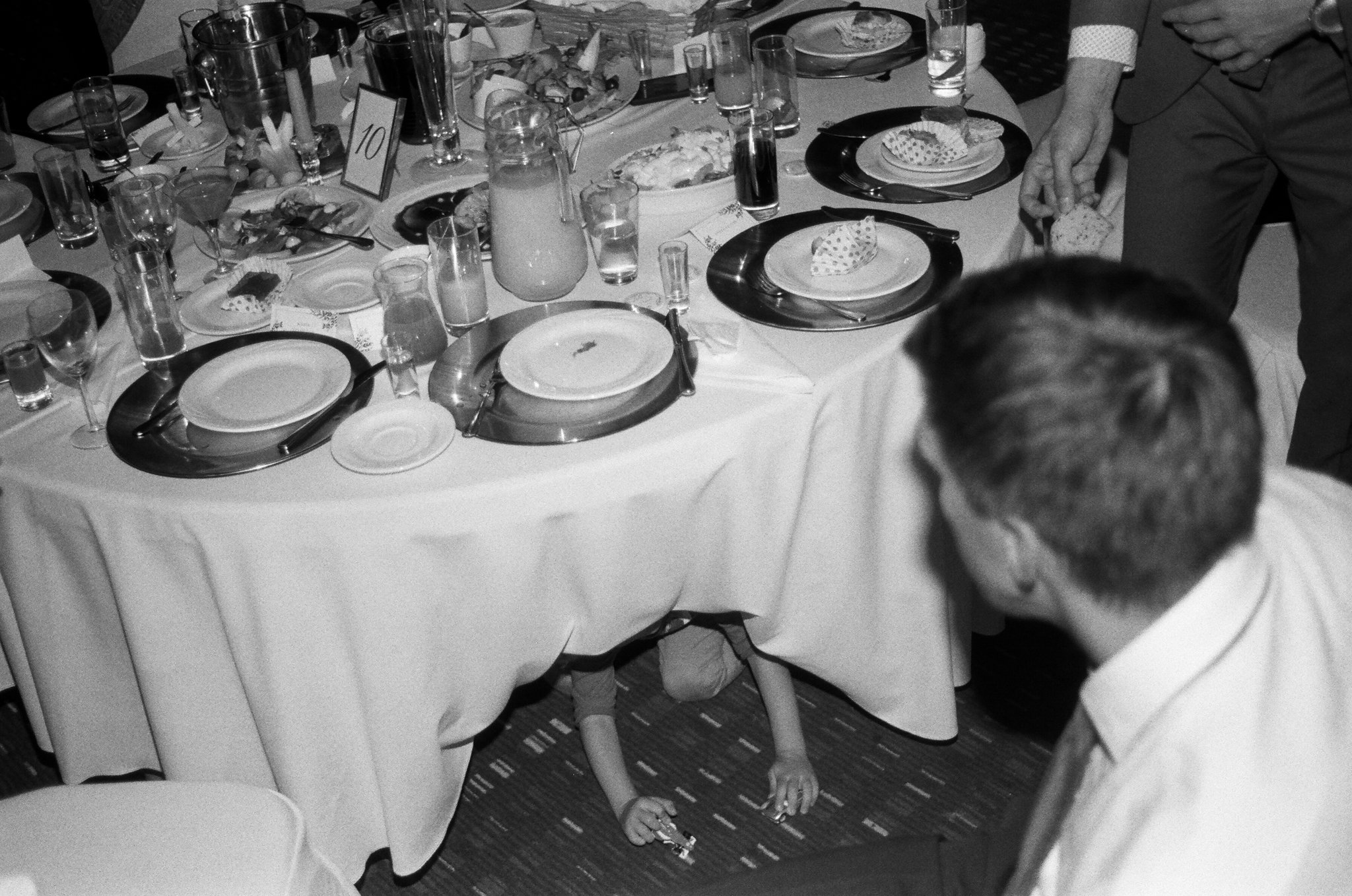 A round banquet table set with plates, glasses, and utensils, with a soup tureen and various food items. A person is seen sitting at the table, and a person is standing, reaching for something. A child is under the table, with only their arms and leg