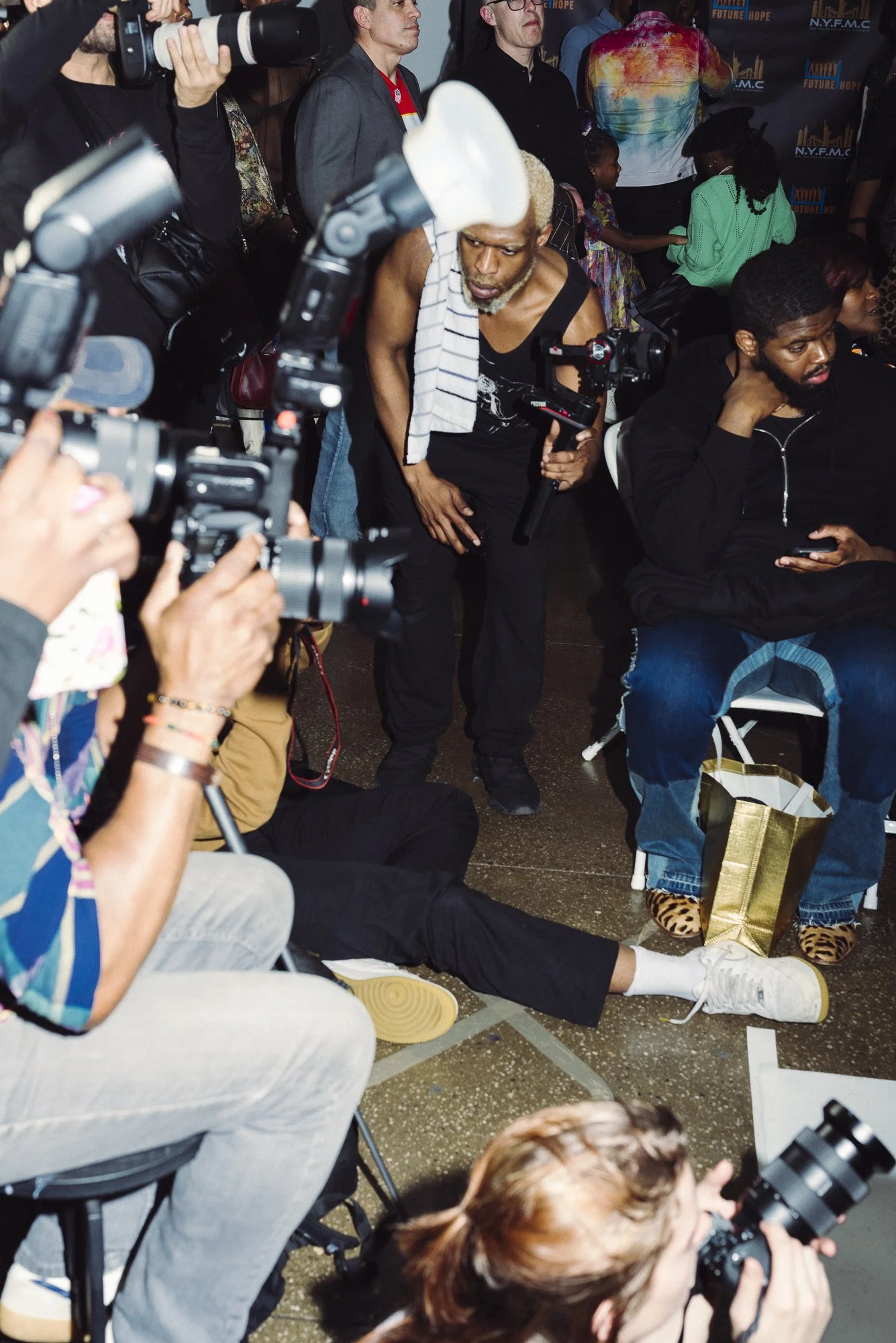 A group of people at an indoor event, some taking photos with professional cameras. An elderly man with gray hair and a towel on his shoulder is standing, holding a camera. Sitting nearby, a man with a phone is looking to his side, and a woman is sit