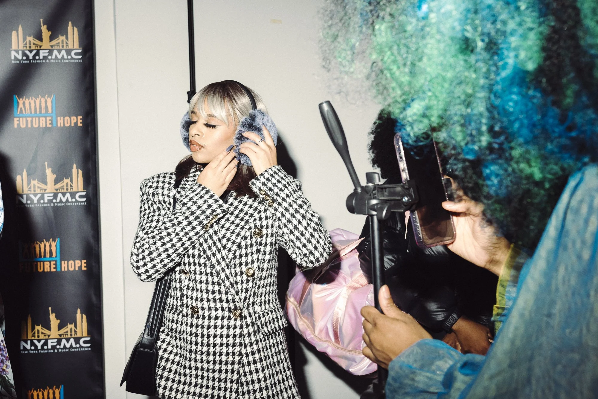 A woman listening to music with headphones, wearing a black and white houndstooth coat, with her eyes closed, at an event with a background displaying the NYFMC logo.