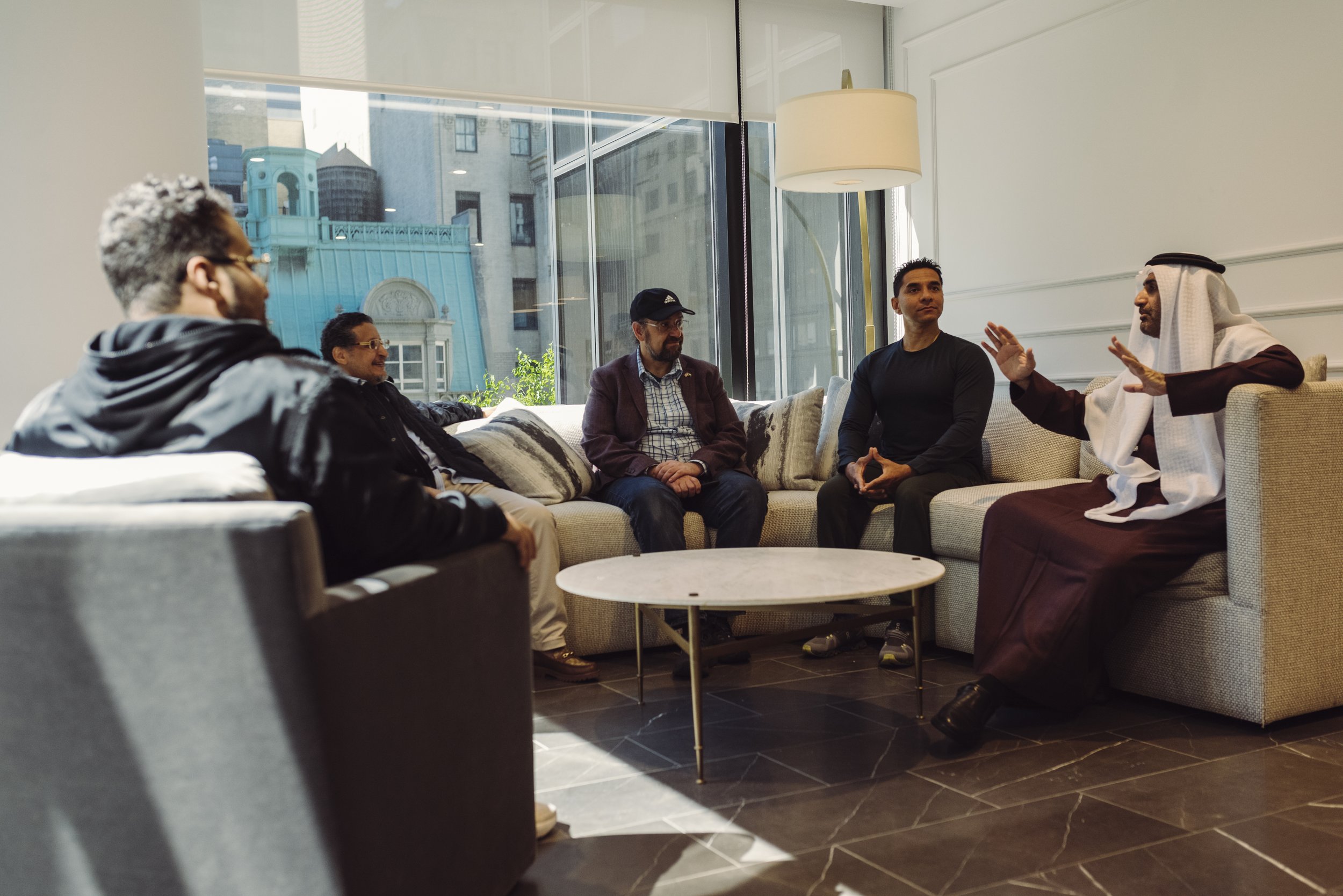Five men sitting in a lounge, engaged in conversation, with a large window behind them showing city buildings.