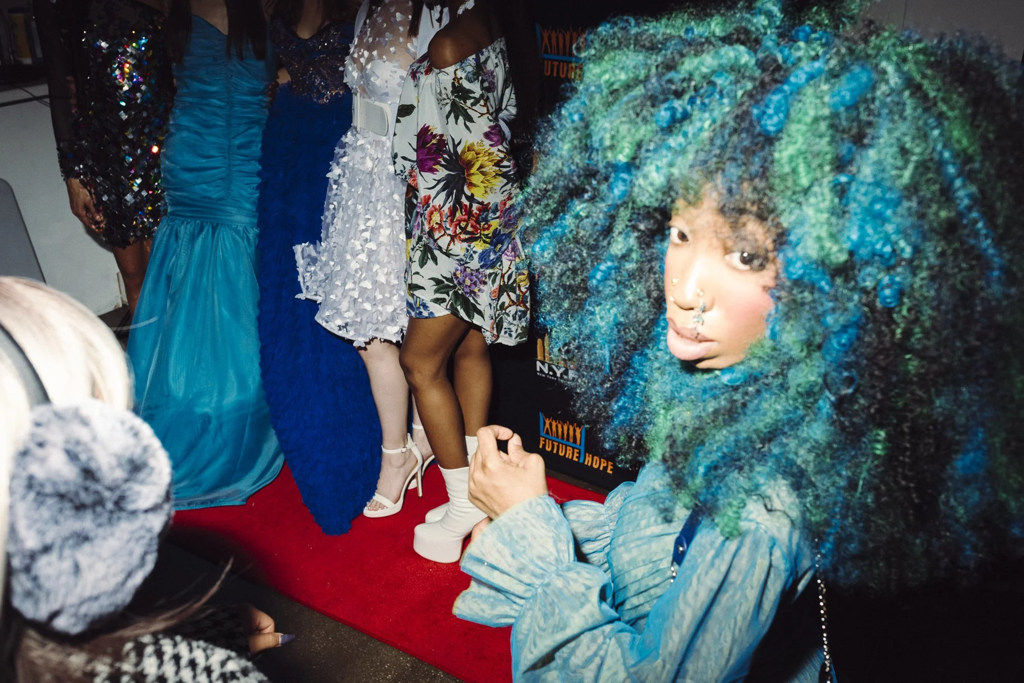A woman with voluminous, curly hair dyed in shades of blue and green, wearing a grayish-blue jacket, posing for the camera at an event with a red carpet, alongside several women dressed in colorful and fashionable dresses.