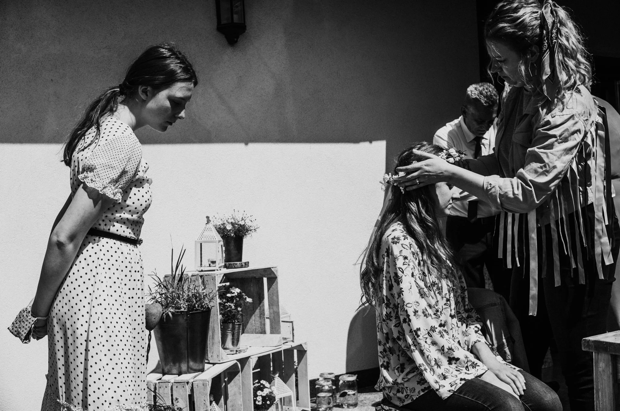 A woman is sitting down with a flower crown being placed on her head by another woman, while a young girl watches. There are plants and decorative items in the background.