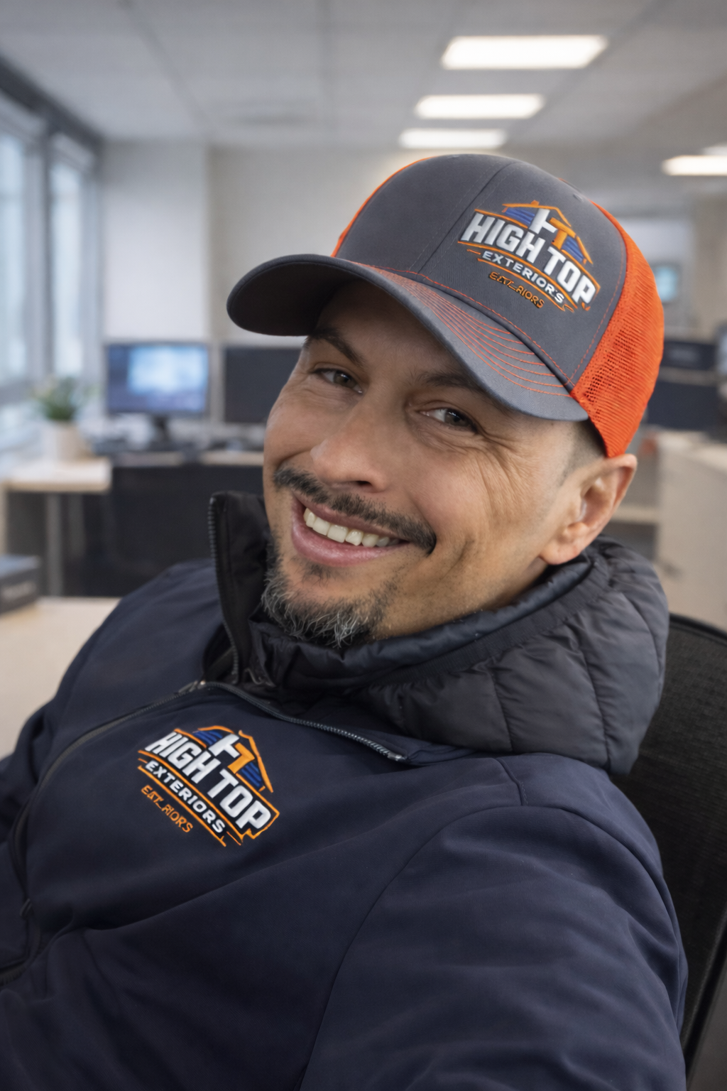 A man smiling in an office wearing a gray and orange baseball cap and a navy jacket, both with the 'High Top Exteriors' logo.