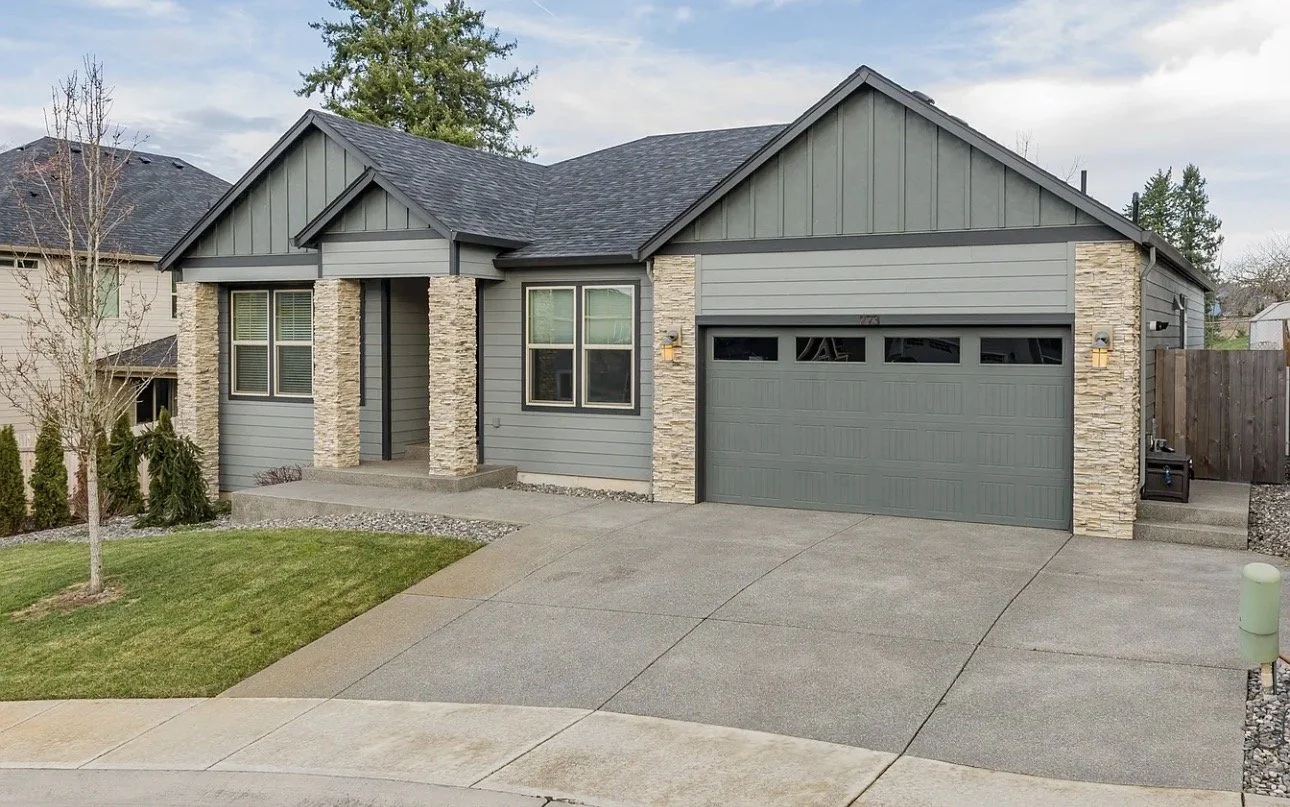 Front view of a modern suburban house with gray siding, stone accents, and an attached garage. The house features a small front porch, manicured lawn, and a tree in the yard.