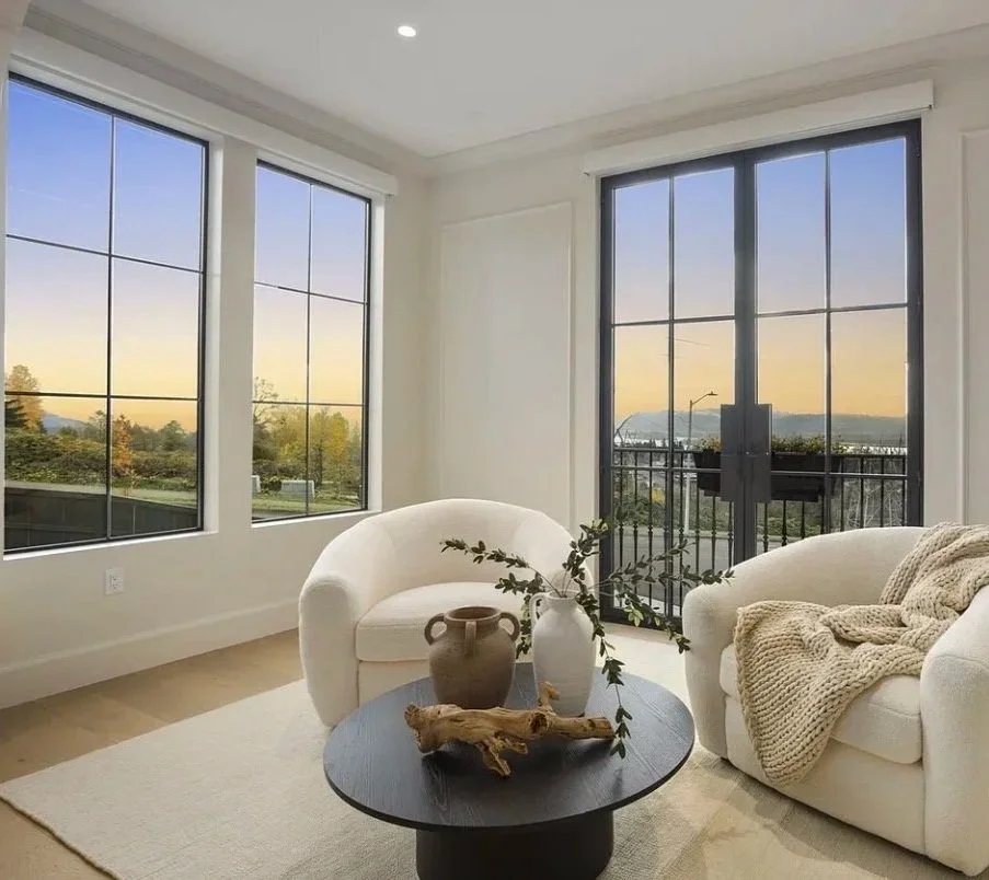 Bright living room with large windows showing a sunset view, white armchairs, a dark round coffee table with decorative vases and a driftwood piece, and a beige knitted blanket.