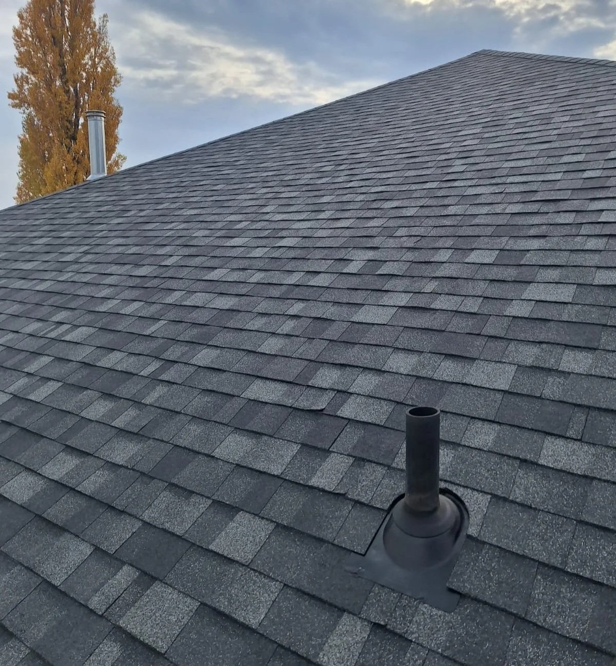 Photo of a gray shingled roof with a vent pipe and a chimney in the background, under a partly cloudy sky.