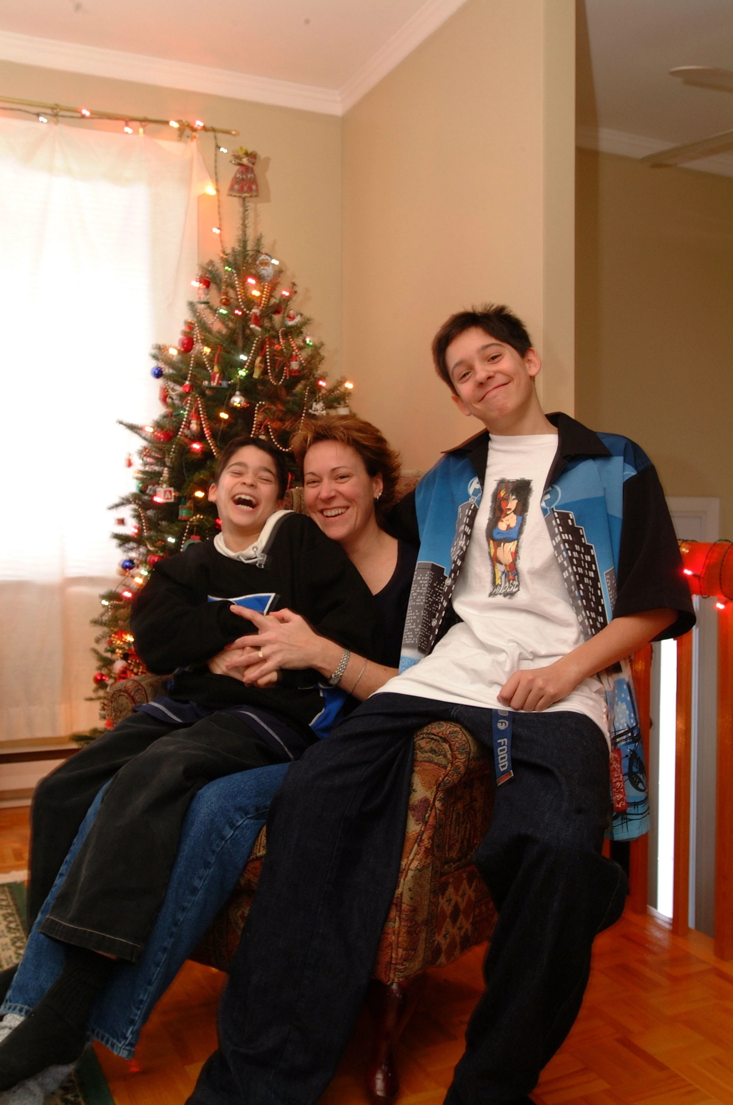 Une femme et deux enfants souriants assis devant un sapin de Noël décoré avec des lumières, des guirlandes et des ornements, dans une pièce éclairée