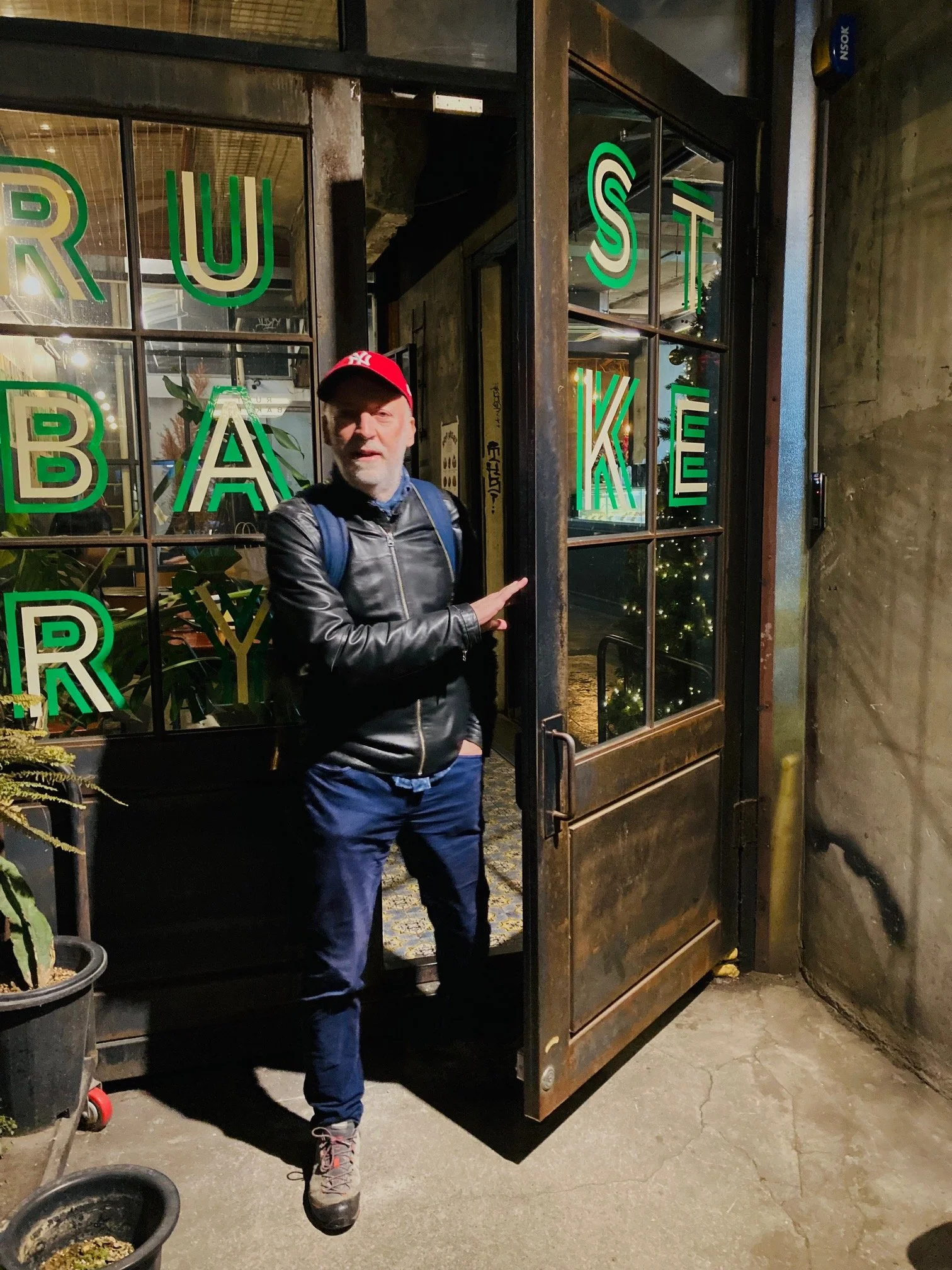 A man standing at the open door of a bar, wearing a black leather jacket, a red baseball cap, blue jeans, and hiking boots. The door has a sign with green letters spelling out 'TRUST BAR'.