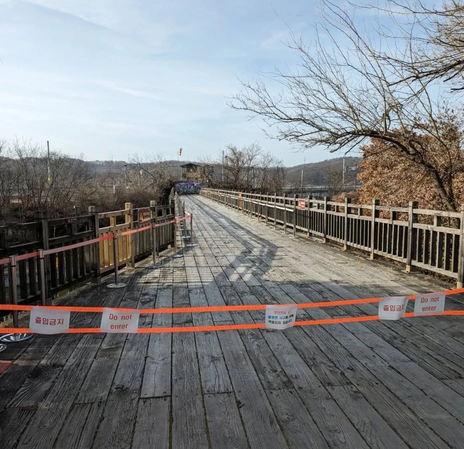 A wooden bridge blocked off with orange and white 'Do not enter' tape, with a railing on each side, over a scenic landscape with bare trees and hills in the background.
