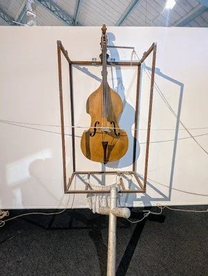A double bass inside a geometric metal frame, with shadows cast on a white gallery wall.