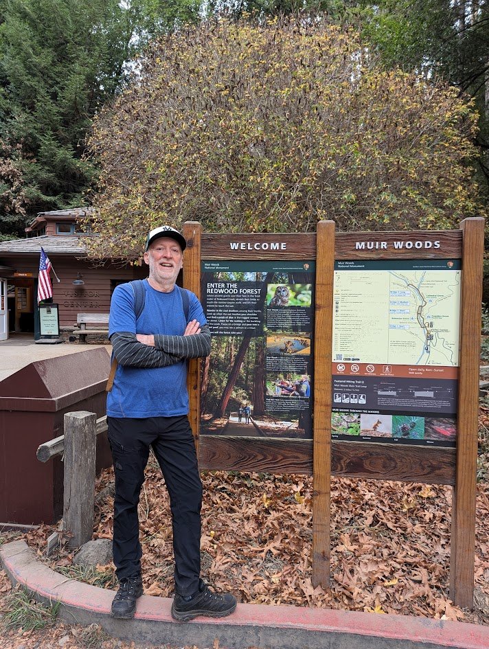 A man standing beside park information boards at Muir Woods, surrounded by fallen autumn leaves and tall trees, with a wooden building and American flag in the background.