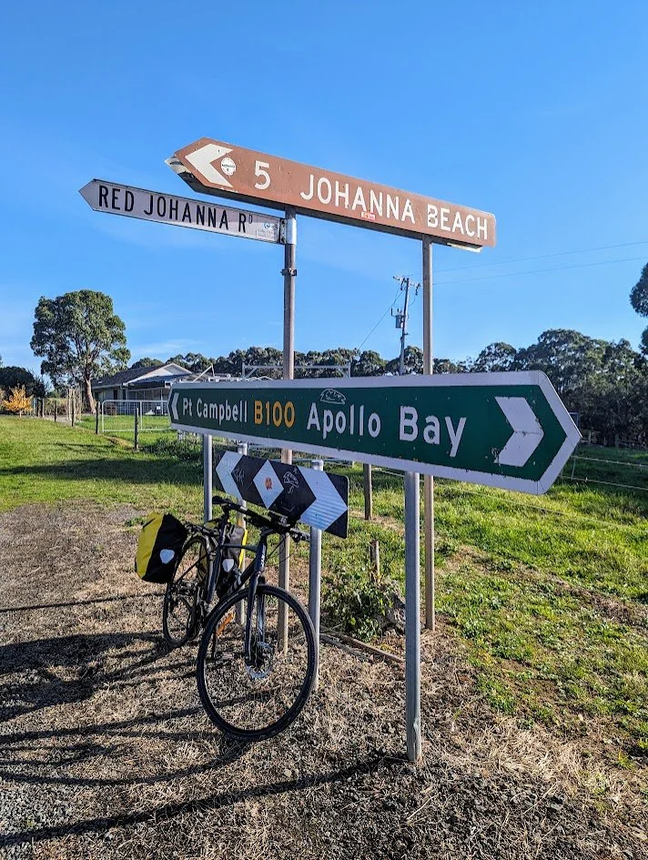 Multiple road signs indicating directions to Johanna Beach, Apollo Bay, and nearby streets, with a bicycle leaning against one sign, on a sunny day with trees and a house in the background.