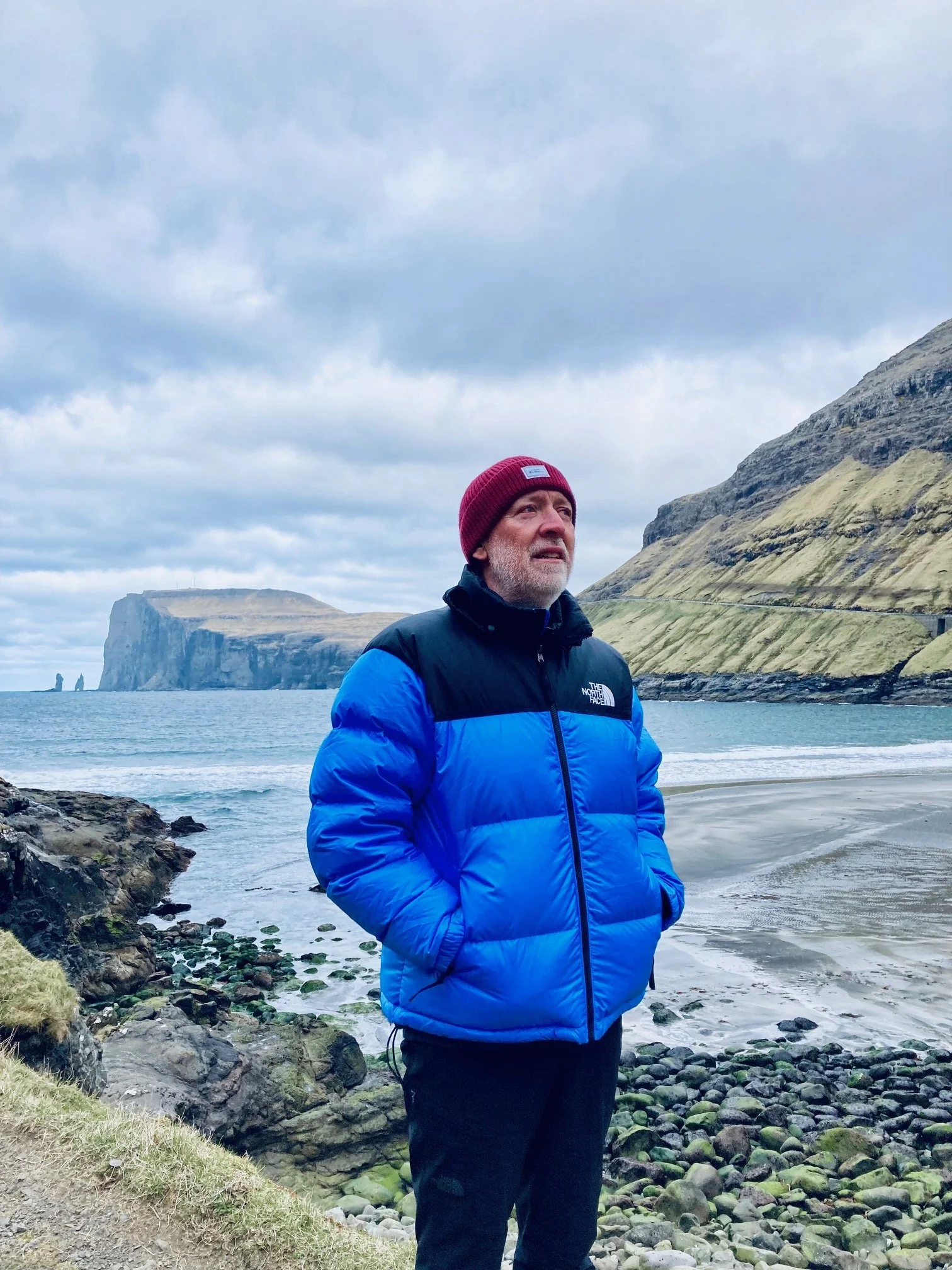 A man wearing a red beanie and a blue and black puffy jacket stands on a rocky beach with cliffs and ocean in the background.