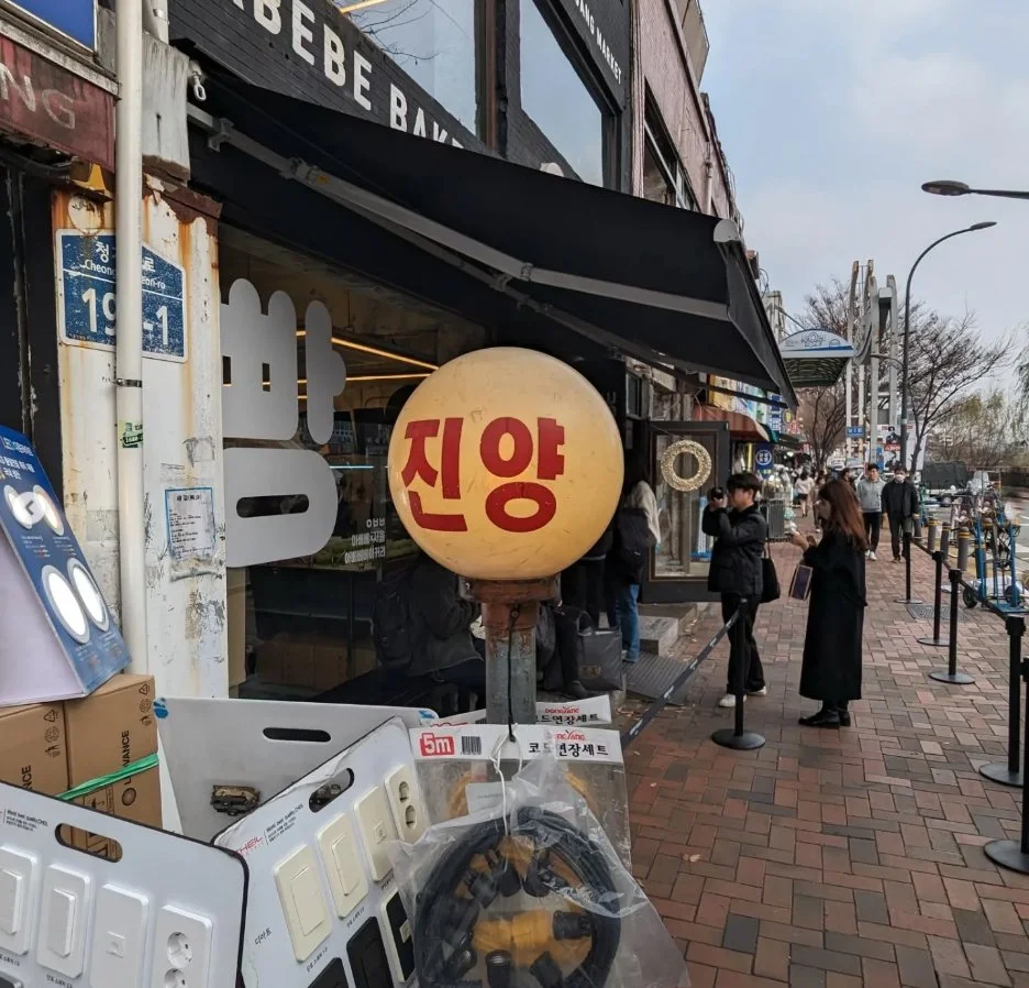 Street scene in South Korea with people standing outside a shop. The shop has Korean signs and a round white lantern with red Korean characters. Various electrical outlets and tools are displayed outside. The sidewalk is brick, and there are trees and street lamps in the background.