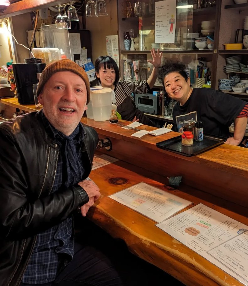 A man in a leather jacket and brown beanie sitting at a wooden bar counter in a restaurant with two smiling women behind the bar, one waving. The background contains shelves with dishes and glasses.