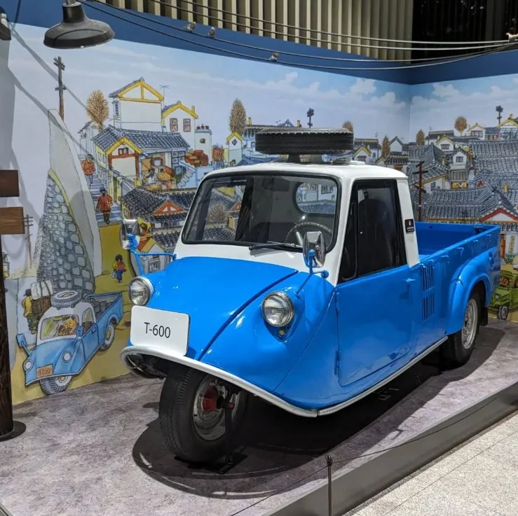 A small vintage three-wheeled vehicle with a blue body and white roof, displayed indoors against a mural of a colorful village scene.