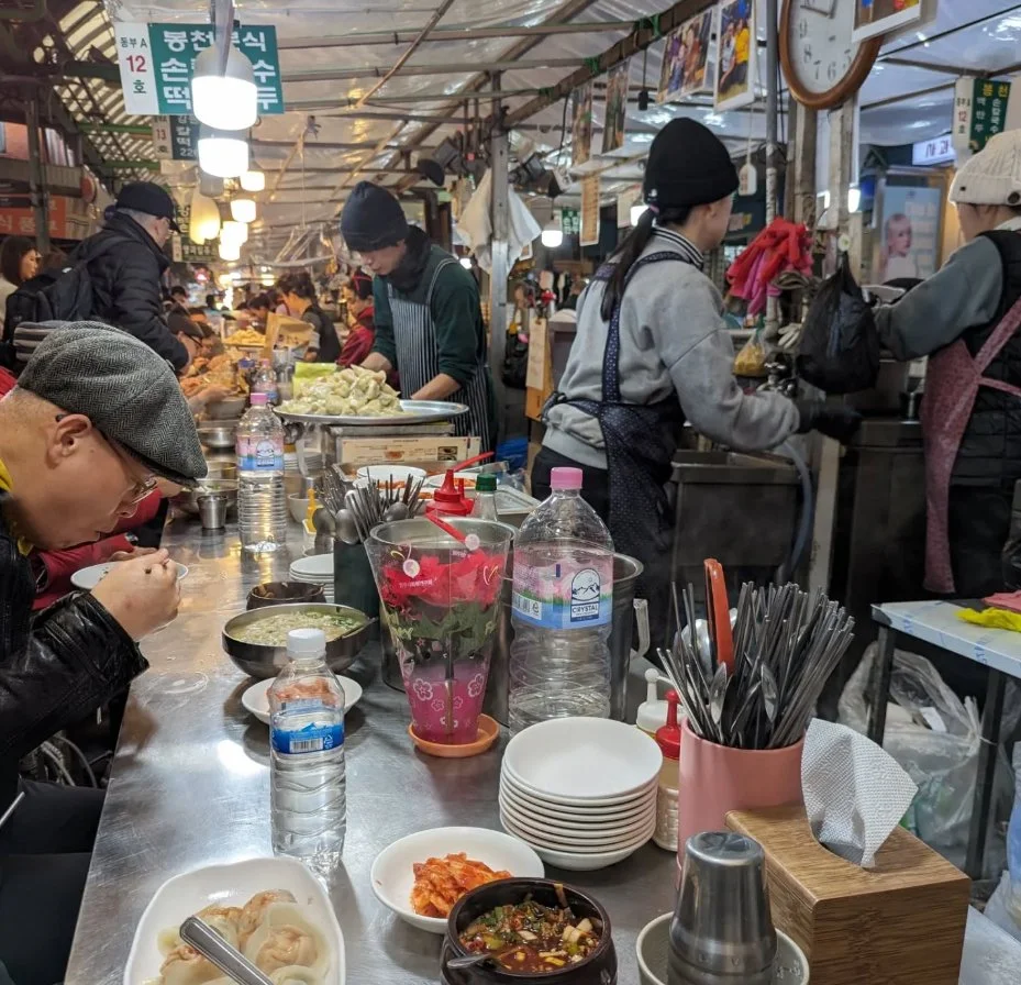People dining at a busy indoor market, some eating soup, other vendors behind the counter preparing food, various dishes, water bottles, and utensils on the table, overhead lighting, and signs in Korean.