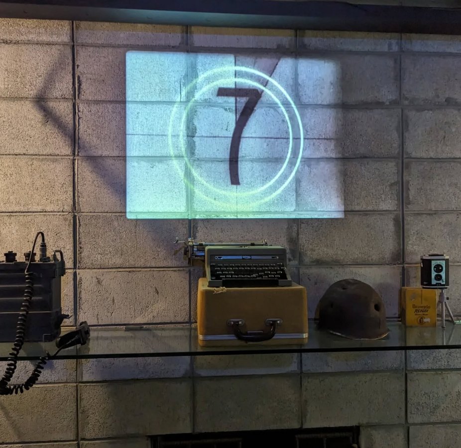 A projection of the number 7 inside a circle on a brick wall, with vintage objects including a typewriter, old helmet, and speaker on a glass shelf below.