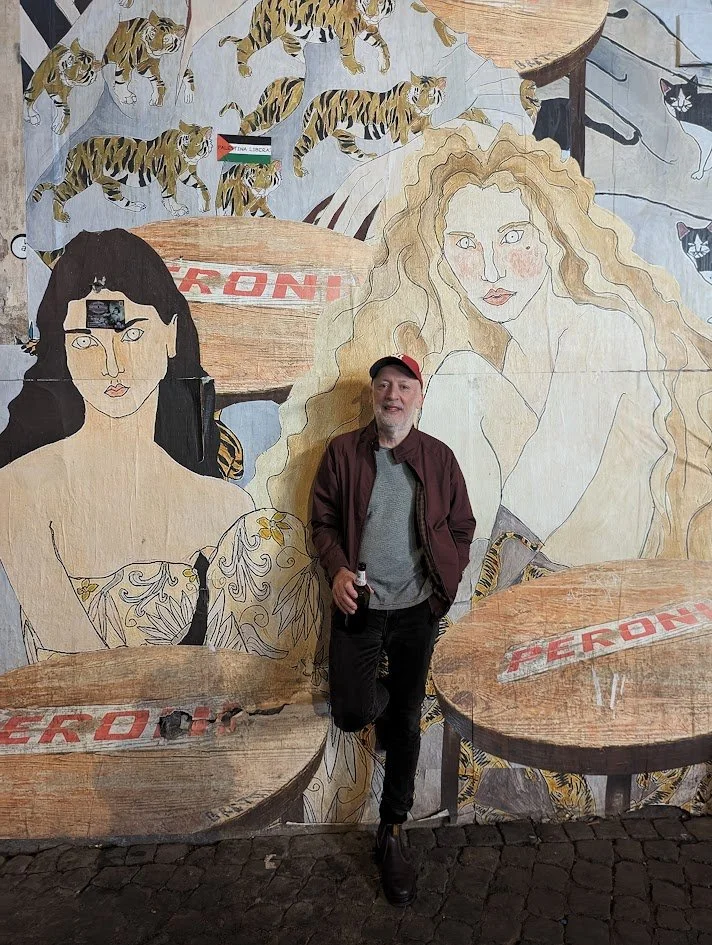 A man stands in front of a large mural featuring stylized portraits of two women and several tigers, with the word 'PERONI' on tables painted on the mural.
