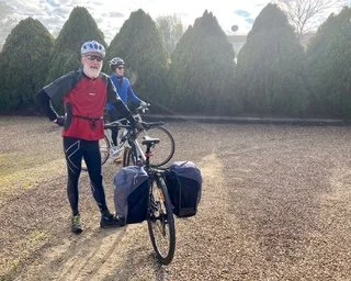 Two cyclists standing outdoors with their bicycles on a grassy area, wearing helmets and athletic clothing, with a line of trees in the background.