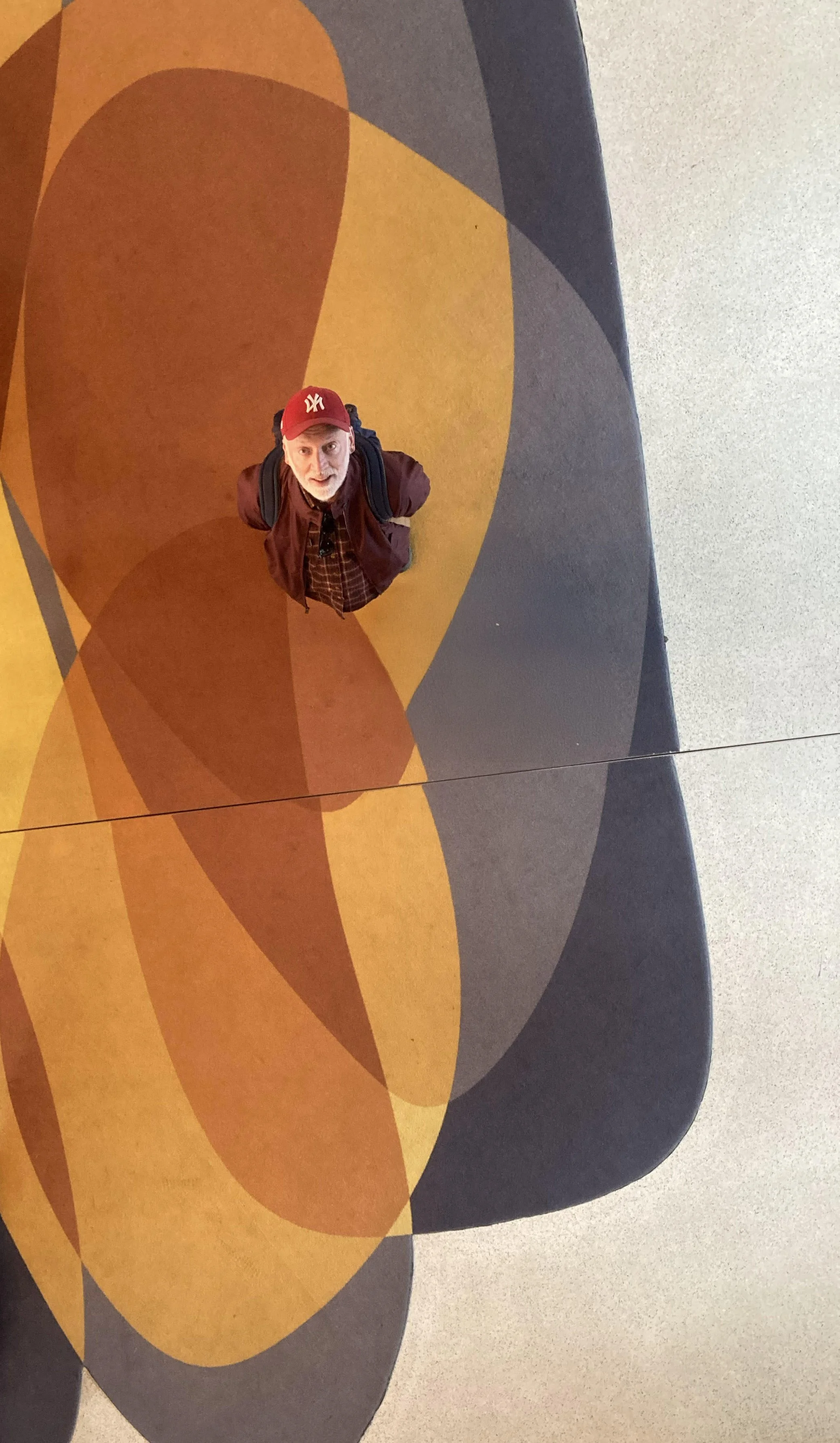 A man with a backpack looking up at the camera in an airport or mall with a colorful geometric design on the floor.