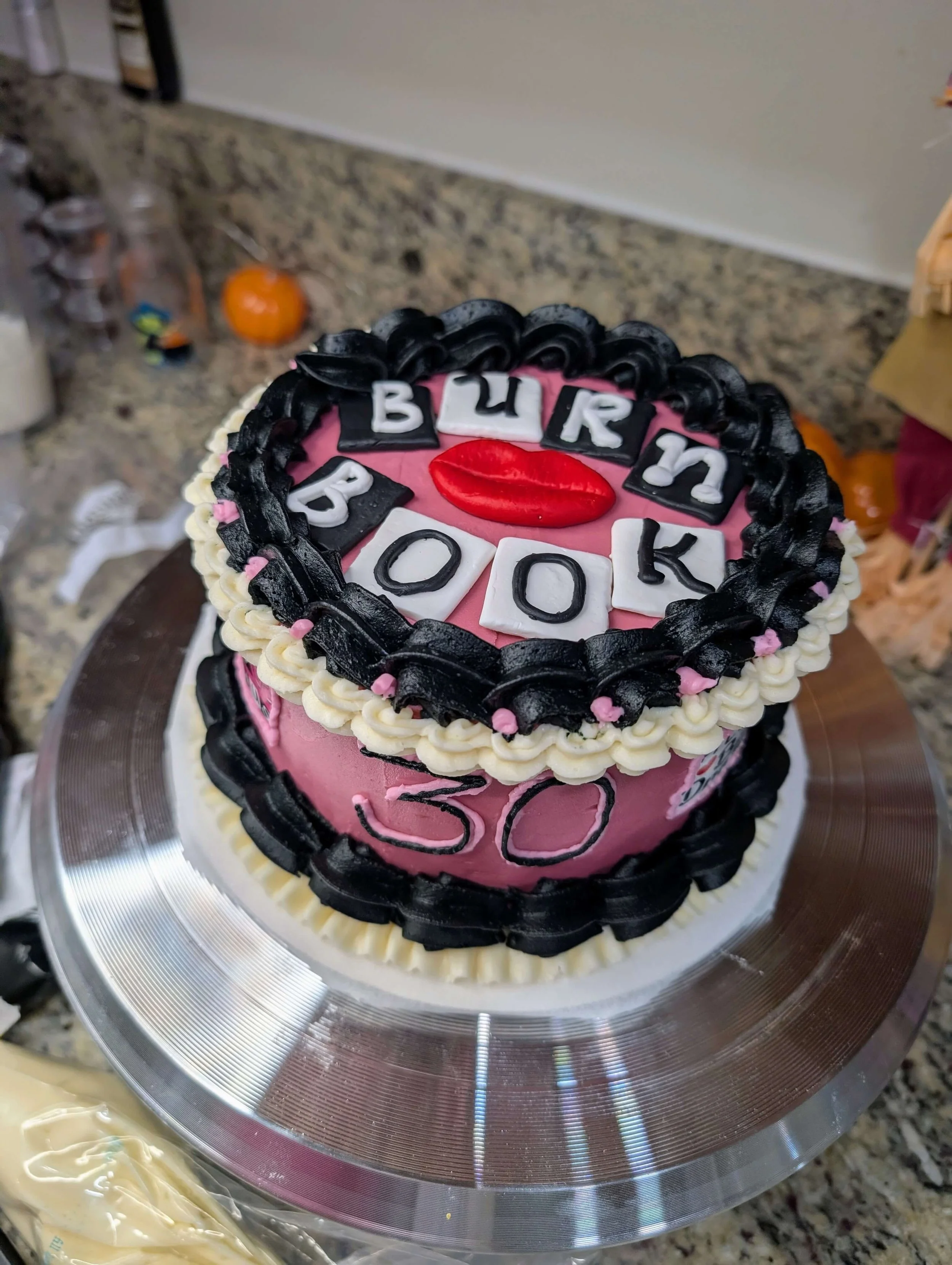 Decorative birthday cake with pink and black icing, topped with a red lips decoration, alphabet tiles spelling 'BURN BOOK', and the number 50 on the side.