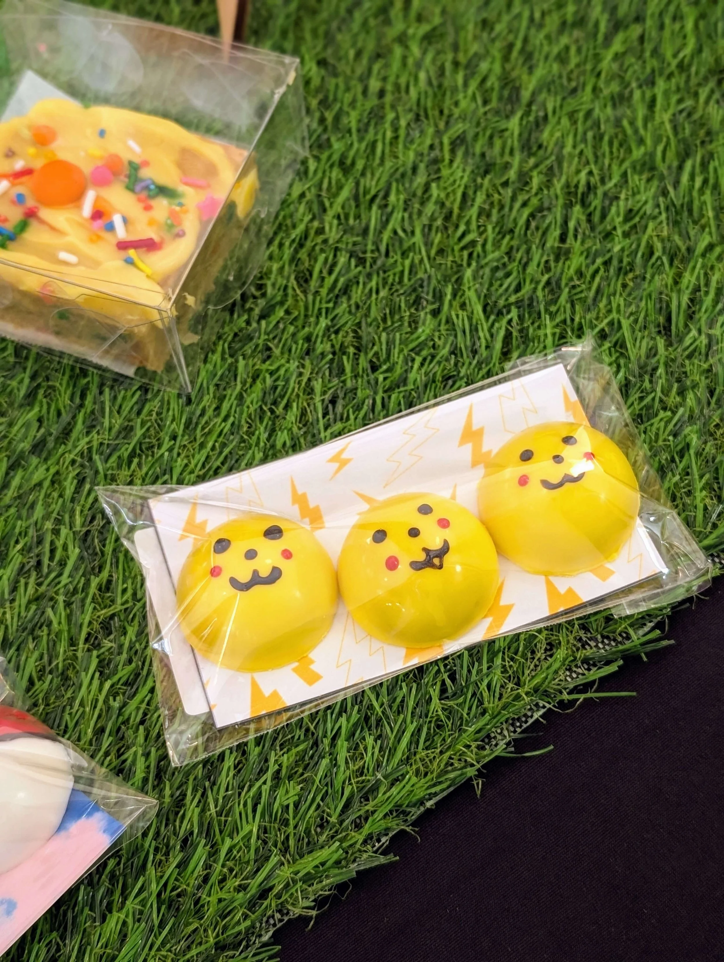 Three round, yellow chocolate bonbons decorated with cute faces resembling Pikachu, arranged in a row inside a clear plastic sleeve with a lightning bolt pattern background, resting on artificial green grass next to other packaged baked treats.