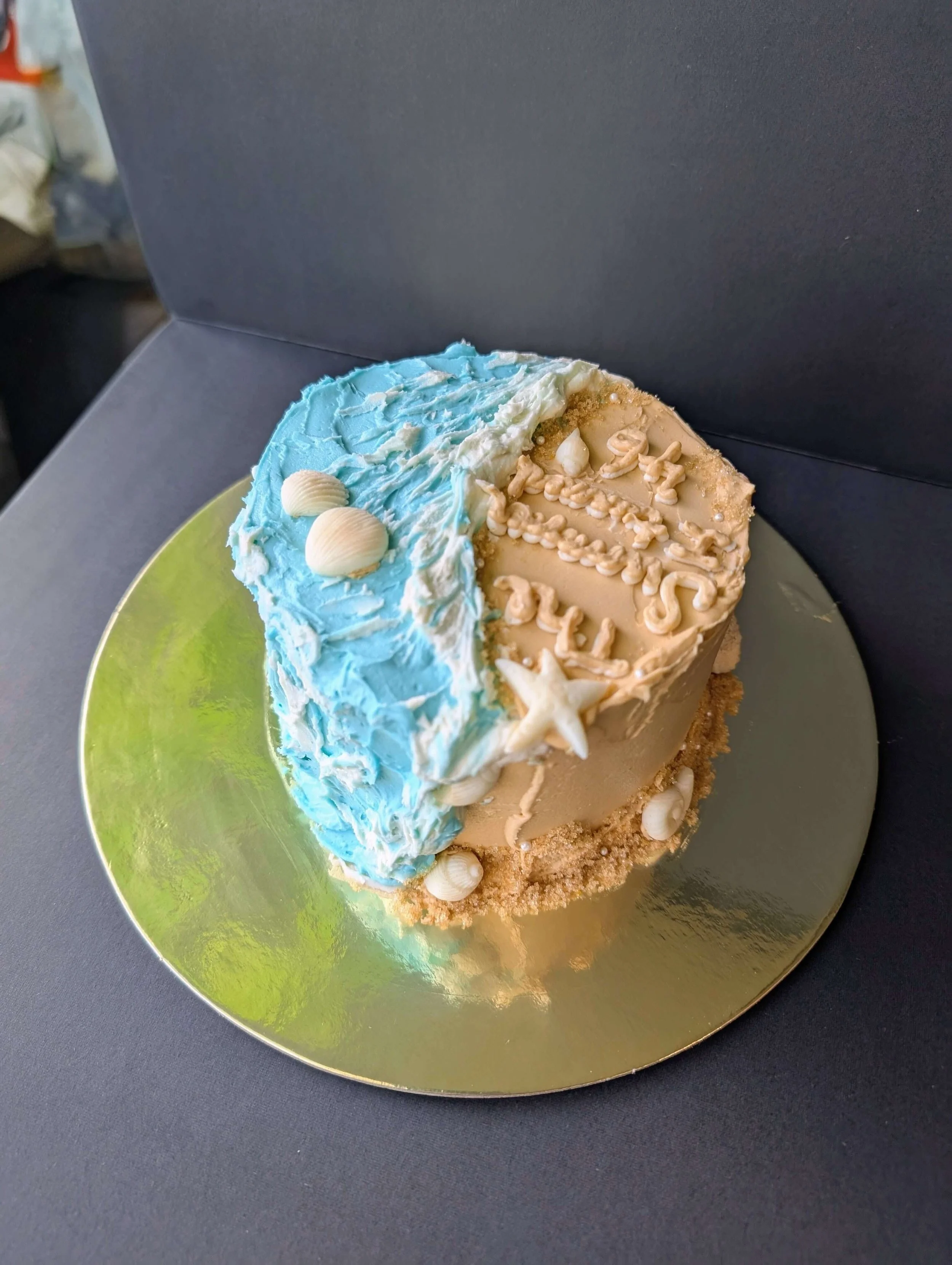 Beach-themed cake decorated with blue and white frosting, seashells, a starfish, and sand-colored details, on a gold cake board.